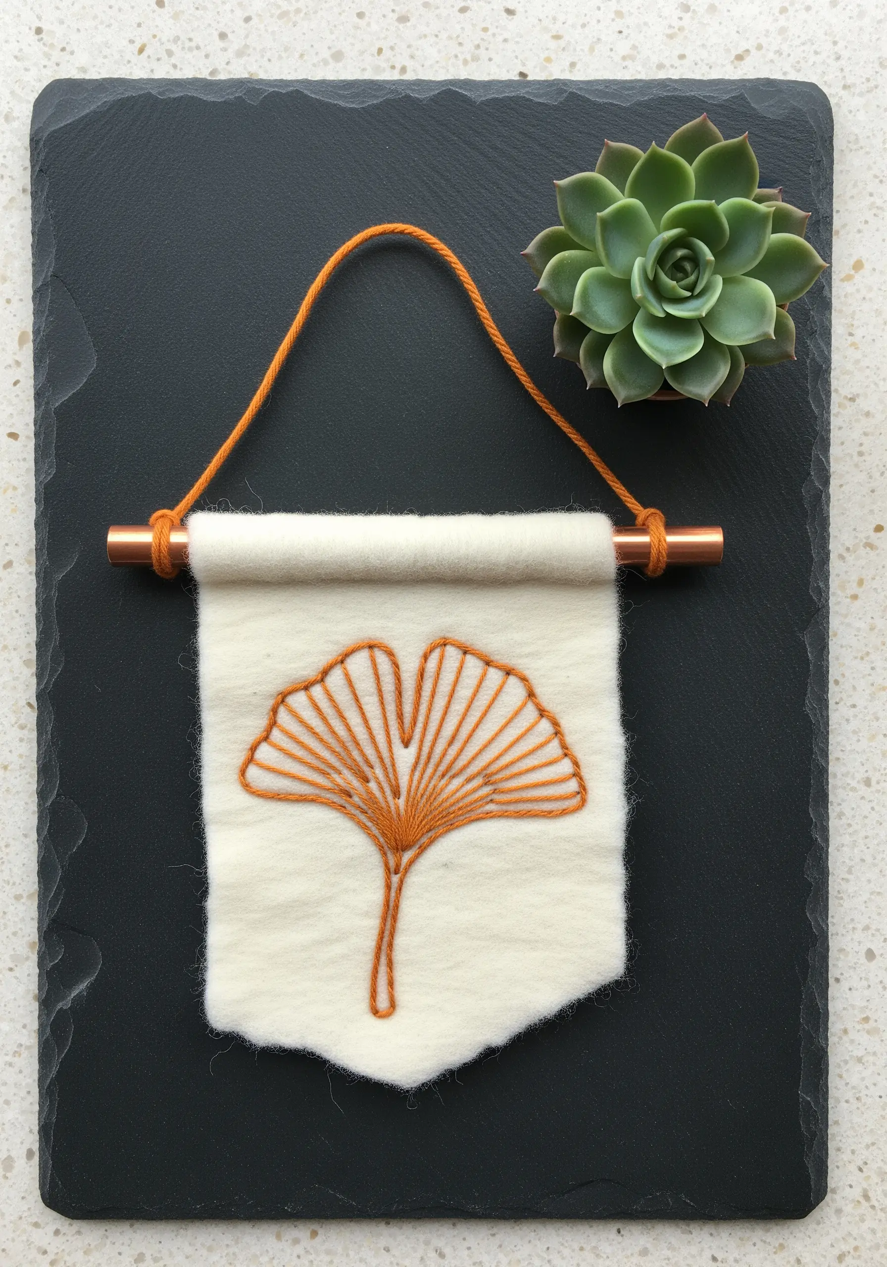 A ginkgo leaf embroidered in orange thread on a cream felt banner with a copper dowel.
