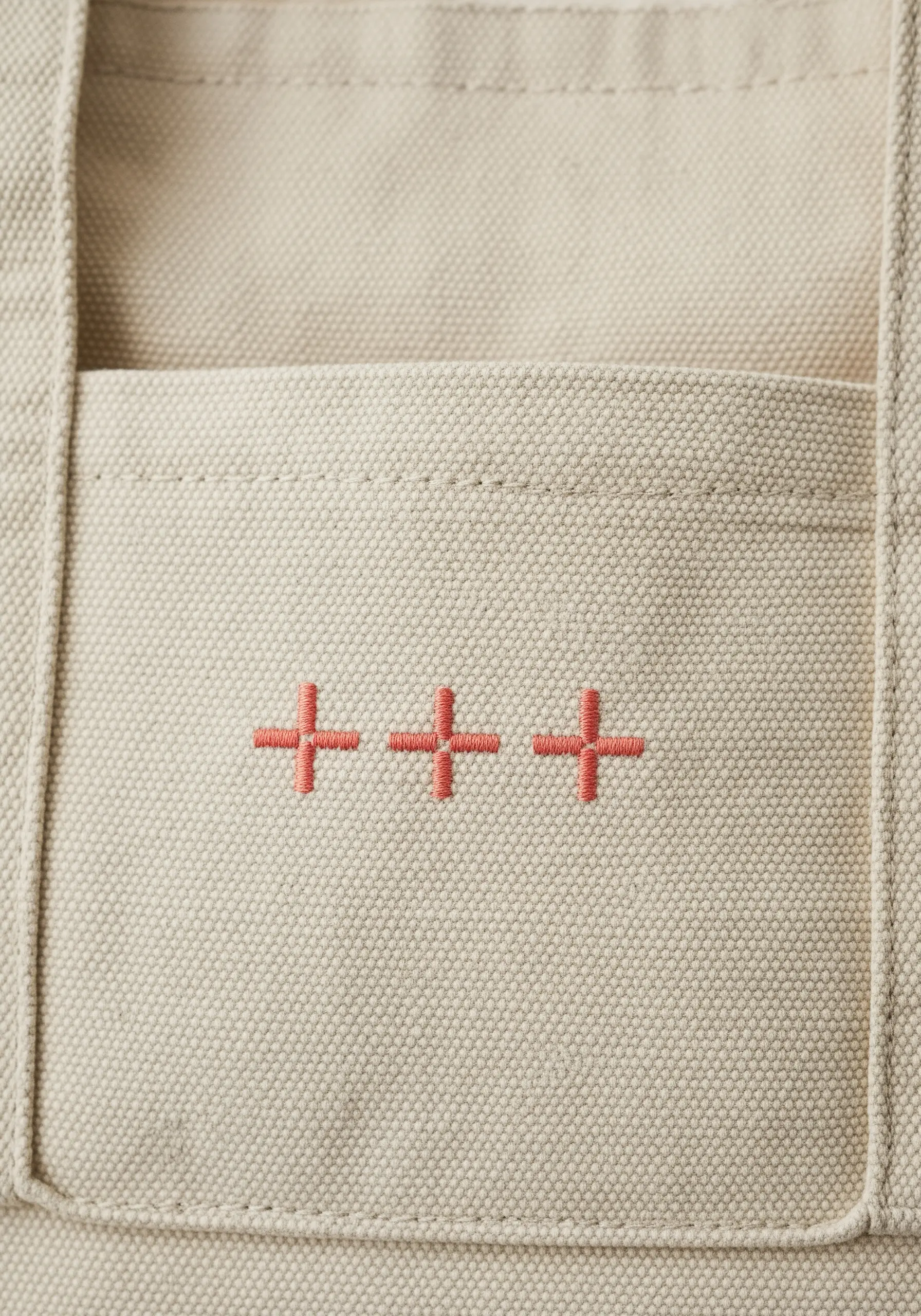 Three small, coral-colored plus signs embroidered in a row on the pocket of a canvas tote bag.