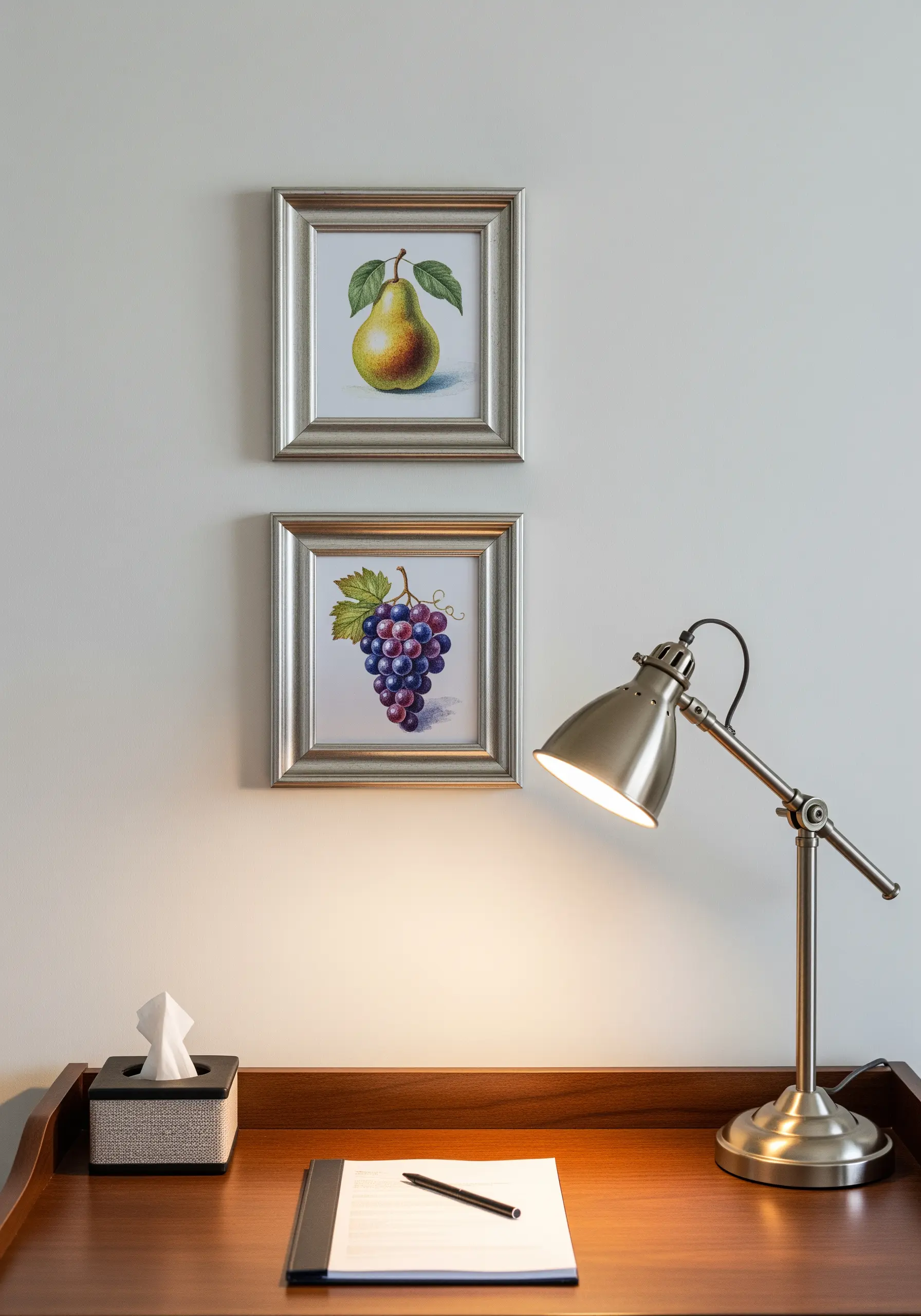 Two silver-framed embroideries of a pear and a bunch of grapes hanging above a desk.