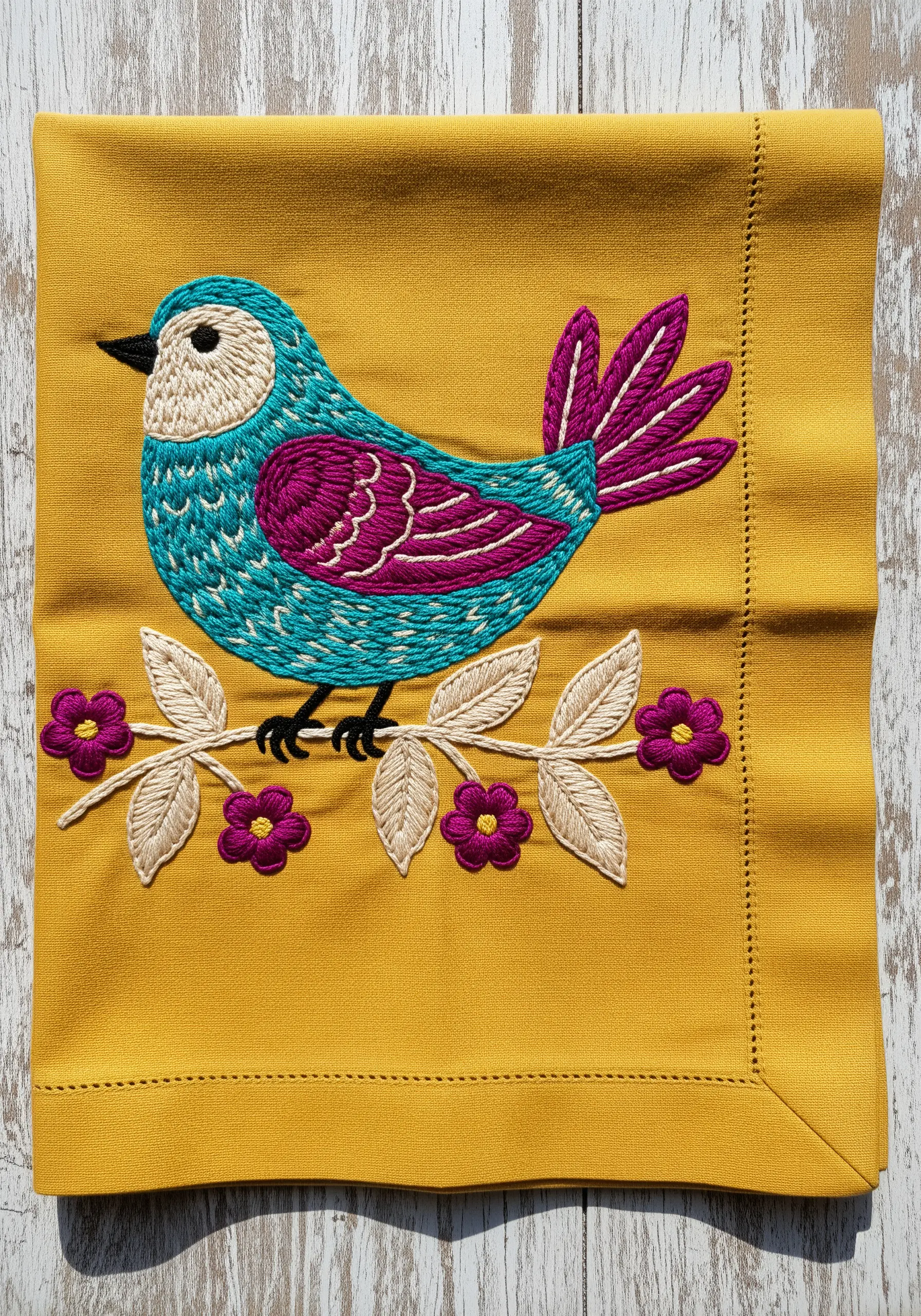 A brightly colored embroidered bird in teal and magenta on a mustard yellow napkin.