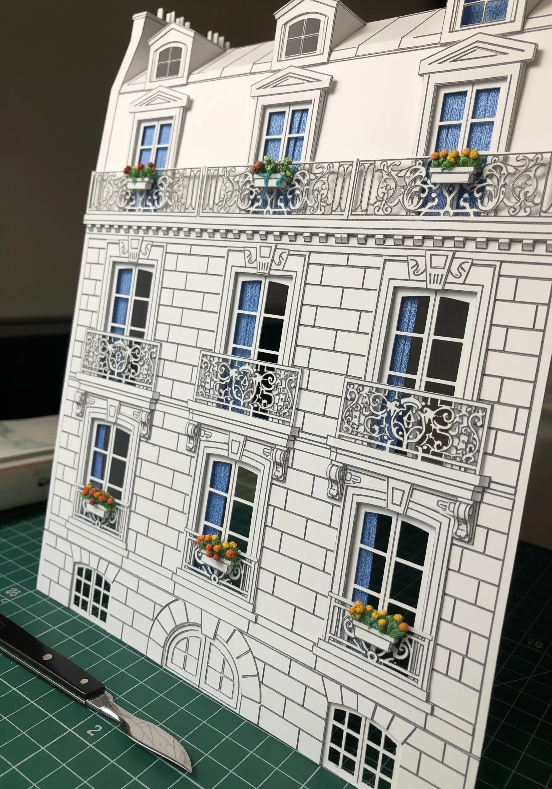 An intricate papercut of a building facade, with blue woven thread visible through the windows.
