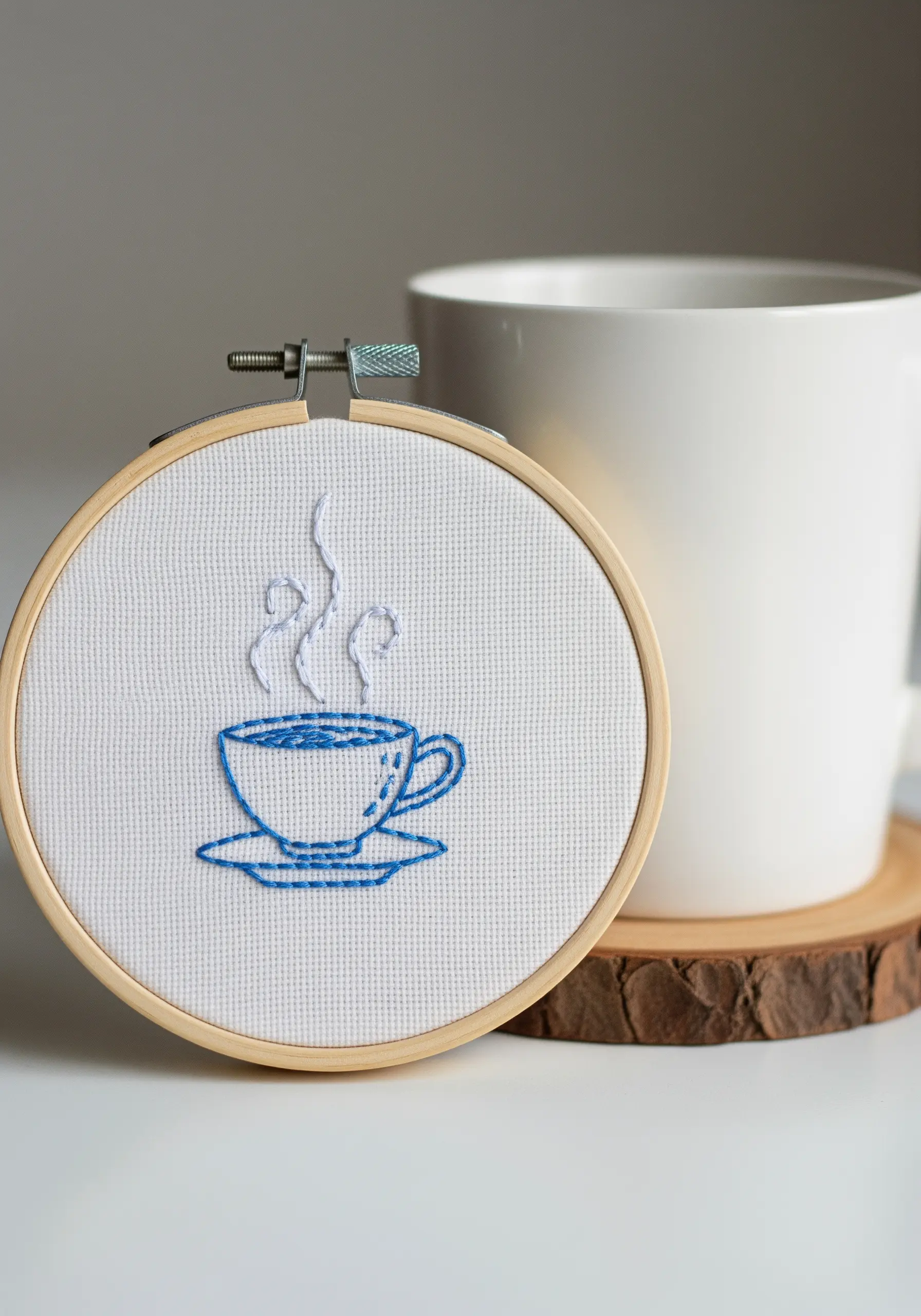 A simple line art embroidery of a steaming coffee cup on white Aida cloth.