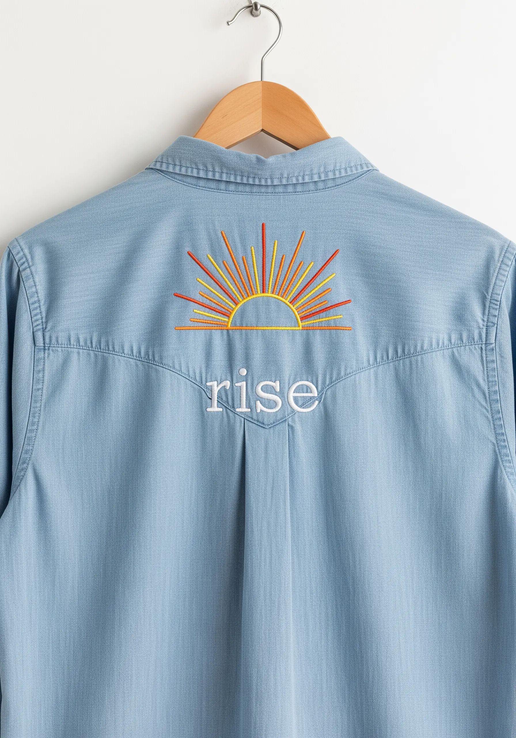 A stylized sunrise with colorful rays embroidered on the back of a chambray shirt above 'rise'