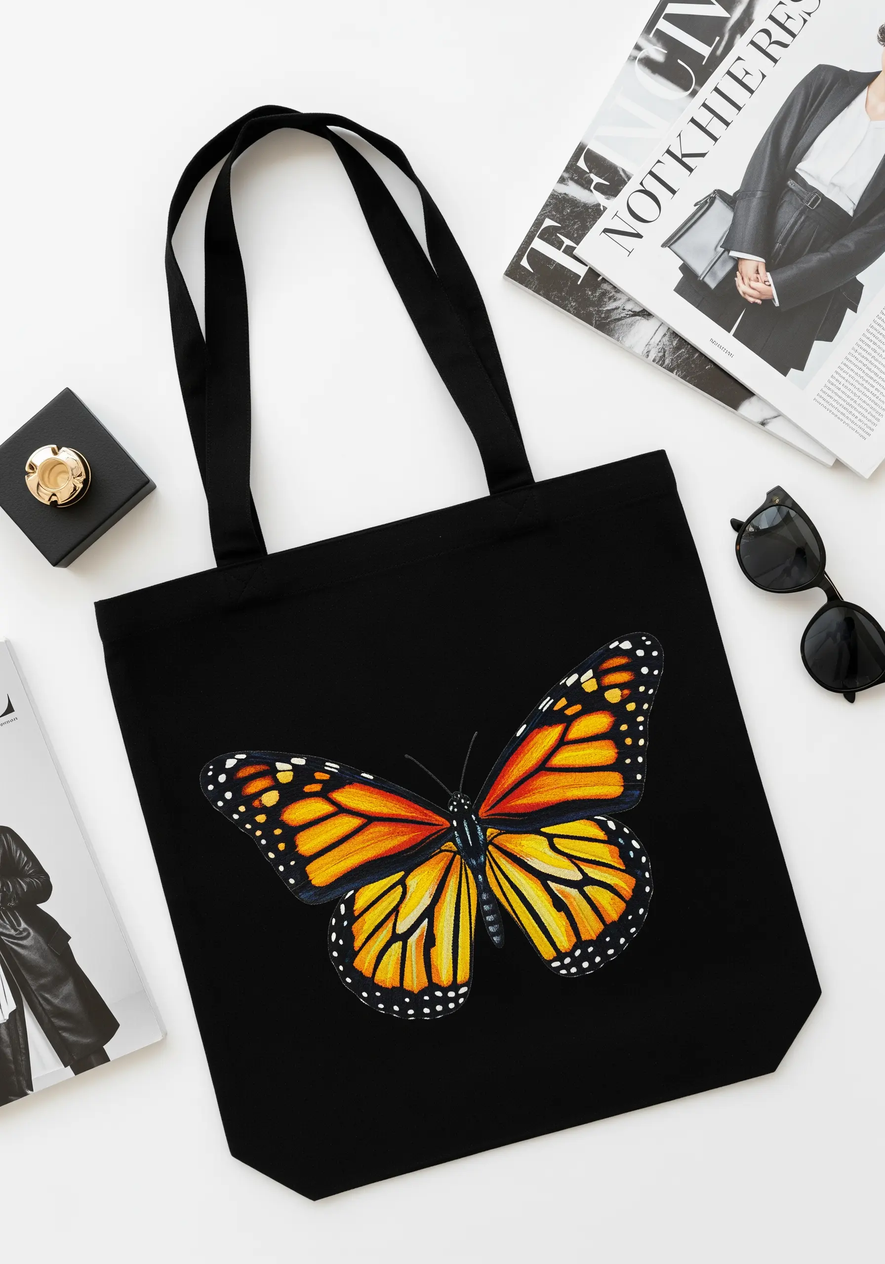 A black canvas tote bag featuring a large, realistically thread-painted Monarch butterfly.