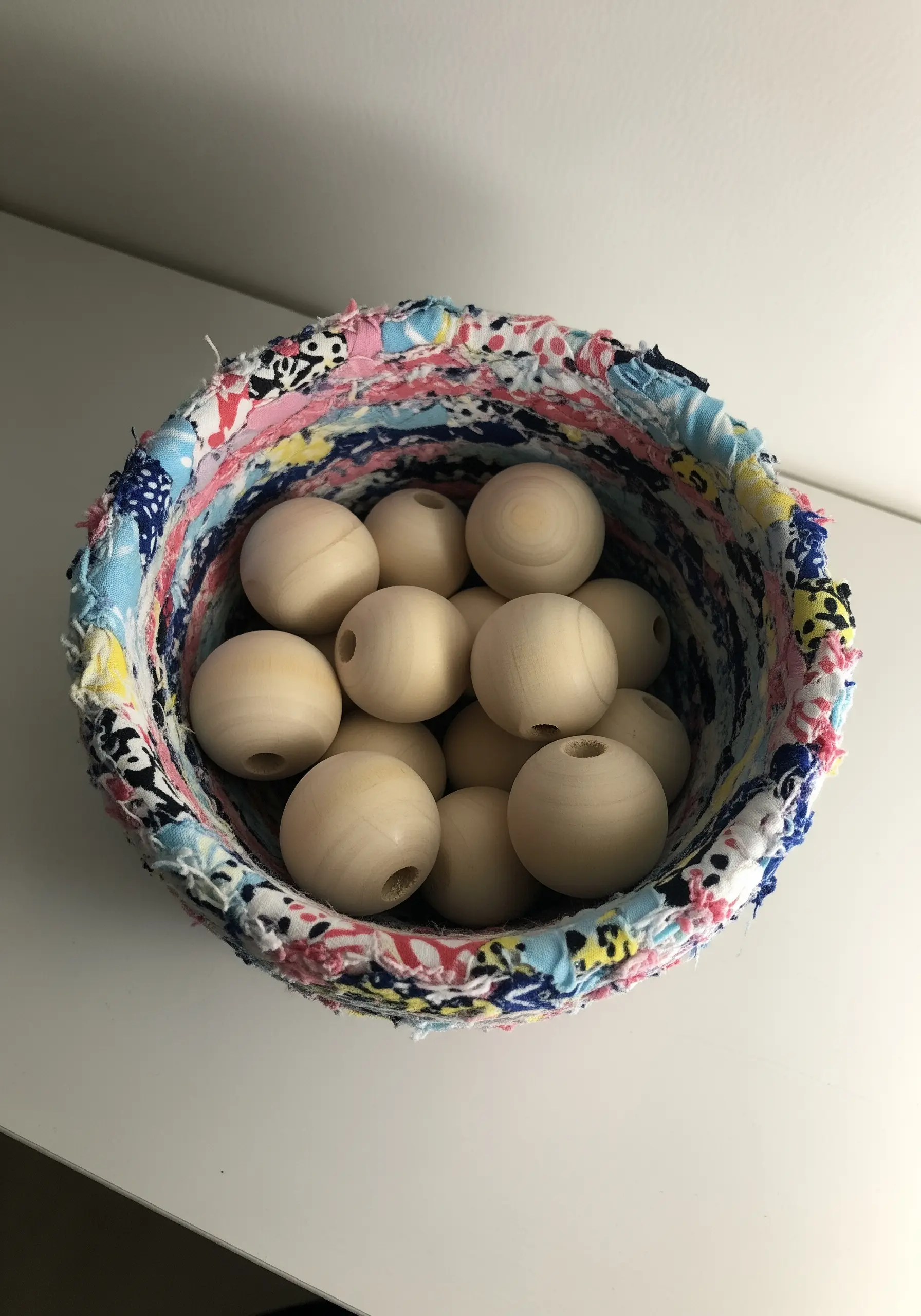 A small, round bowl woven from colorful strips of scrap fabric, filled with wooden beads.