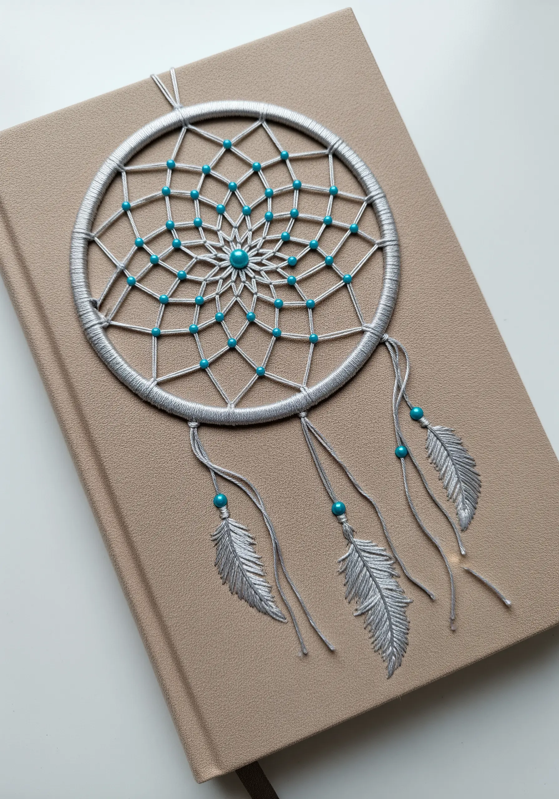 A silver and turquoise dreamcatcher embroidered onto a taupe journal cover.