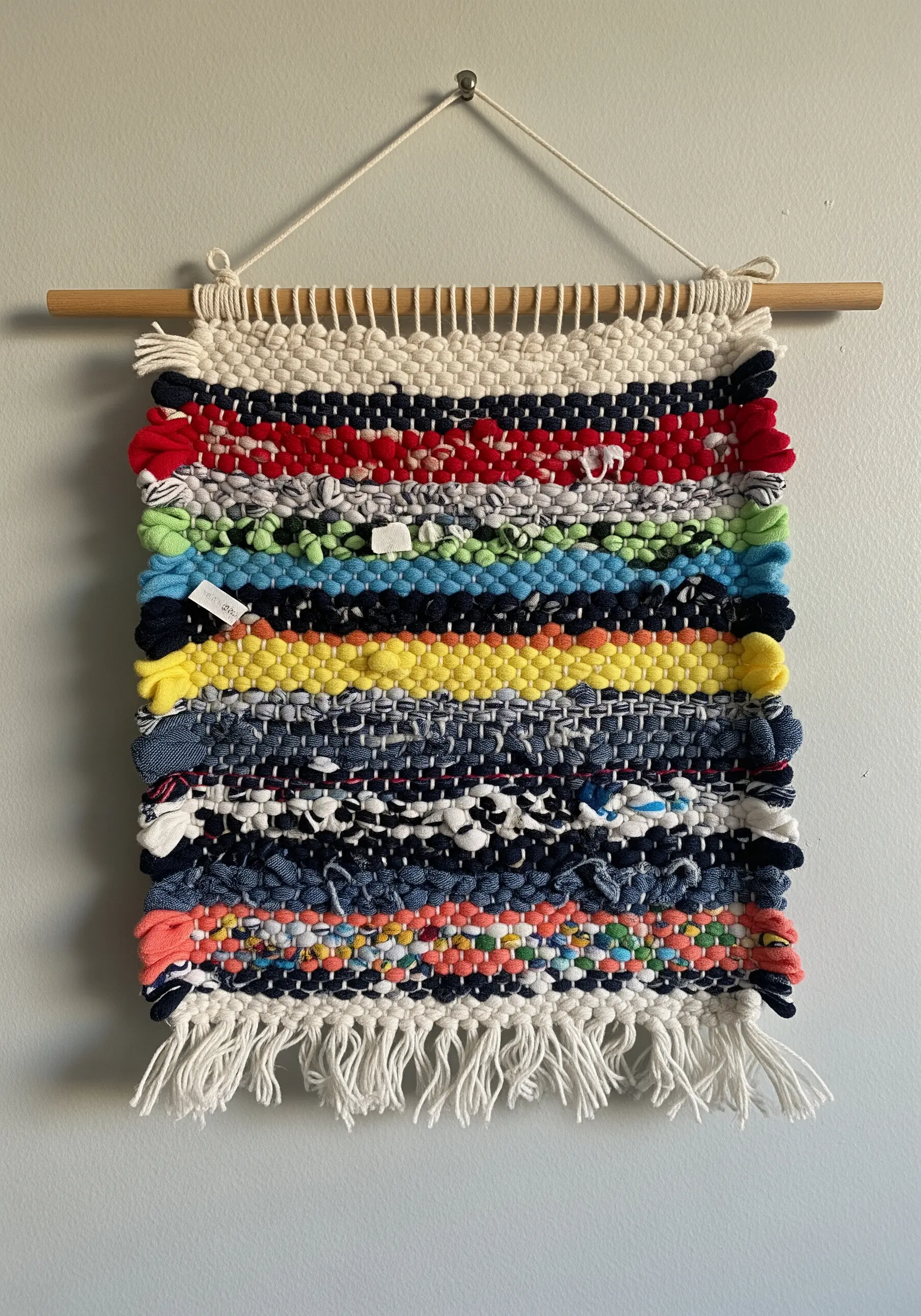 A small, colorful woven wall hanging made from strips of repurposed fabric on a wooden dowel.