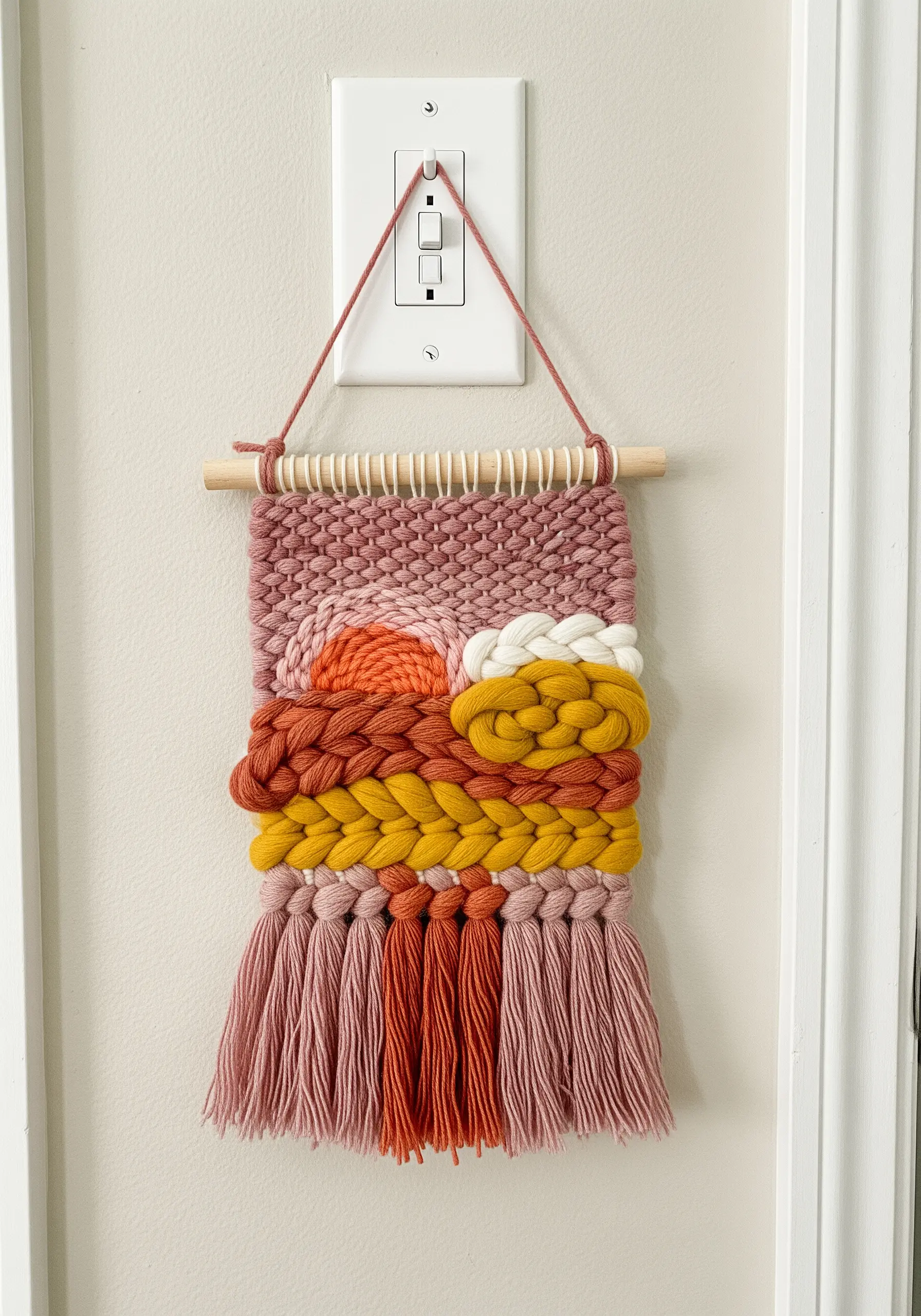 A small woven wall hanging with thick yarn in pink, orange, and yellow hues depicting an abstract sunset.