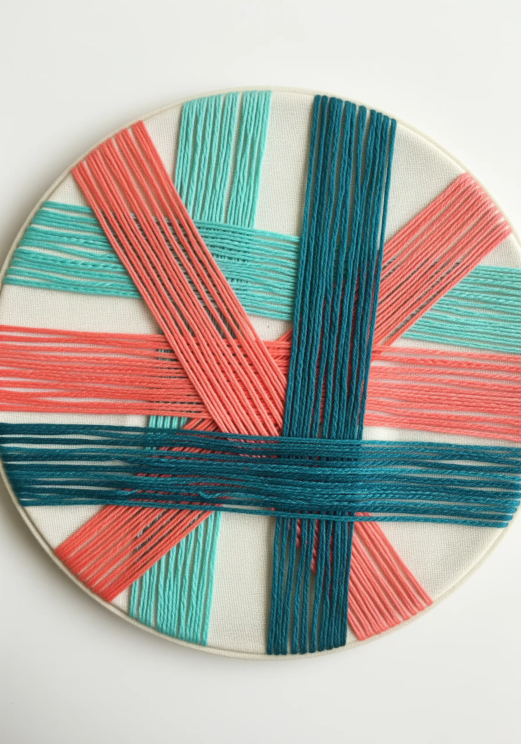 Woven thread art in a hoop, with interlaced bands of coral, teal, and dark blue.