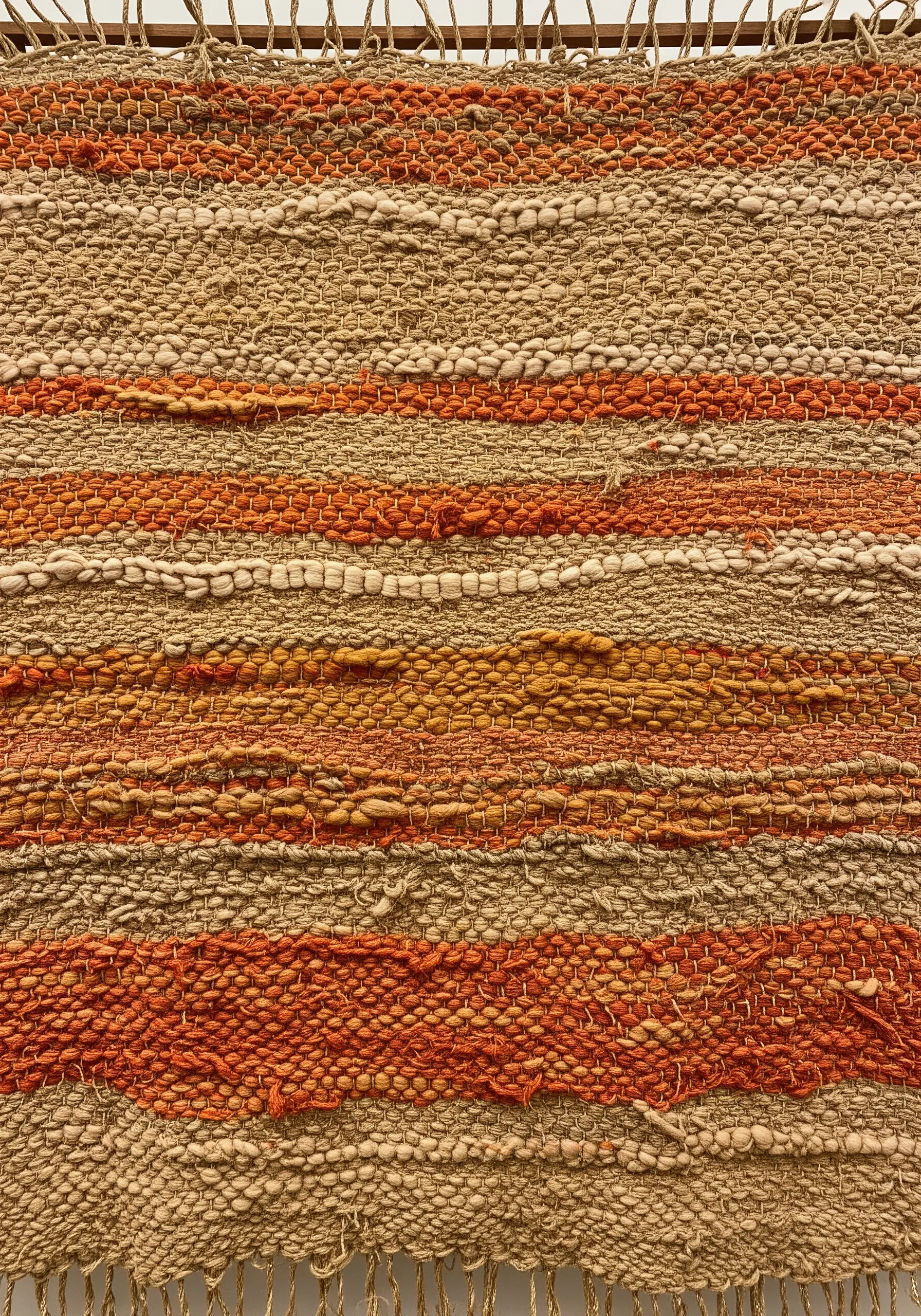 A close-up of a woven textile with thick, textured stripes in shades of beige, orange, and brown.
