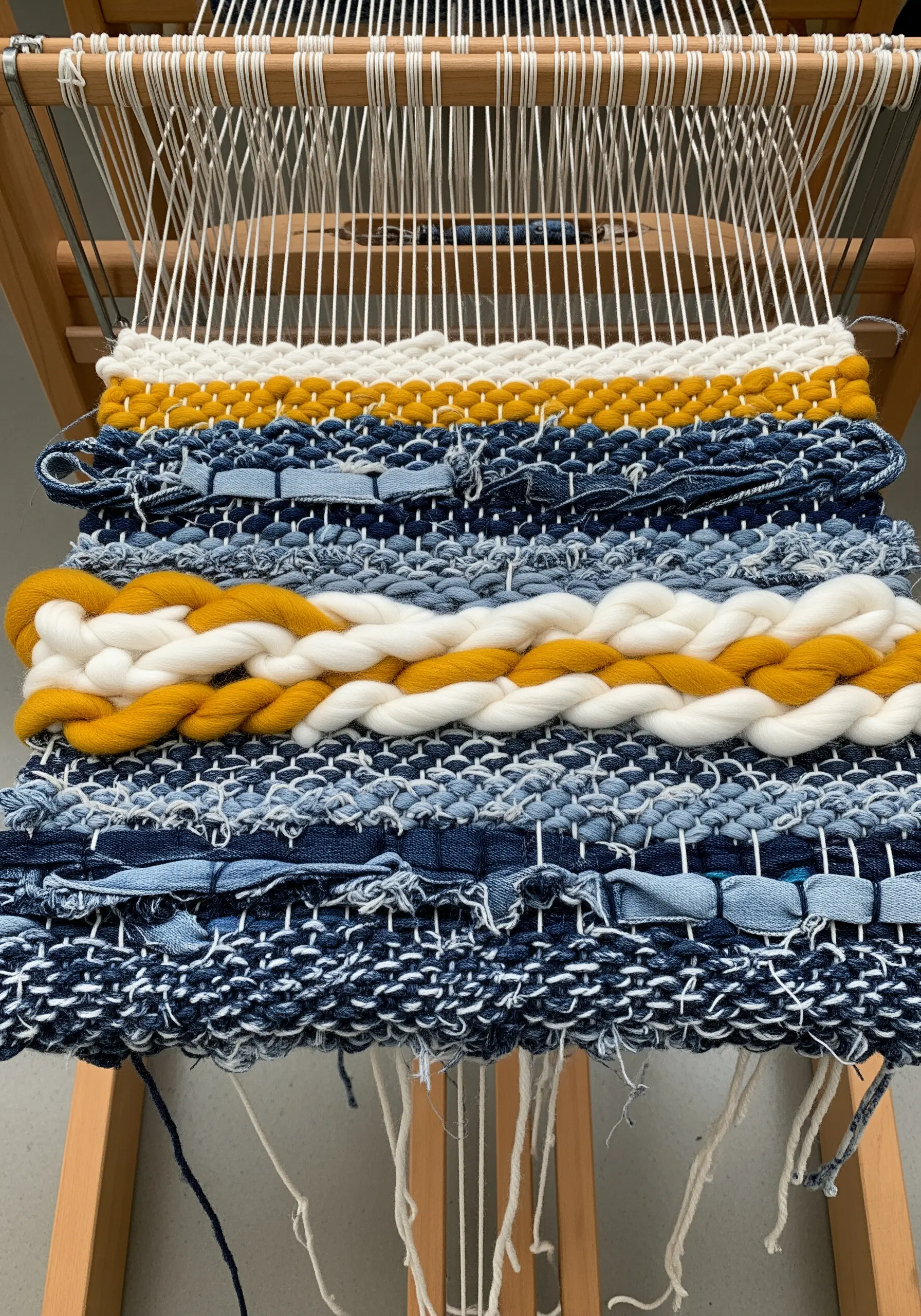 A weaving in progress on a loom, using strips of upcycled blue jeans and chunky yellow yarn.