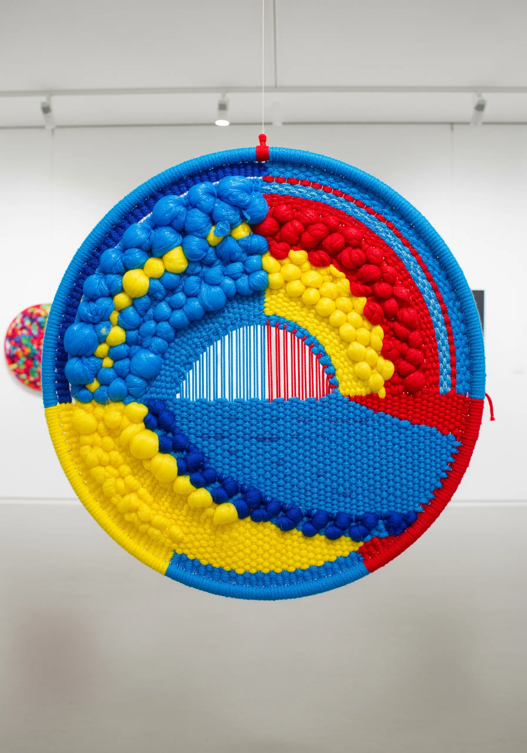 A circular woven wall hanging with bold red, yellow, and blue sections made from recycled plastic yarn.