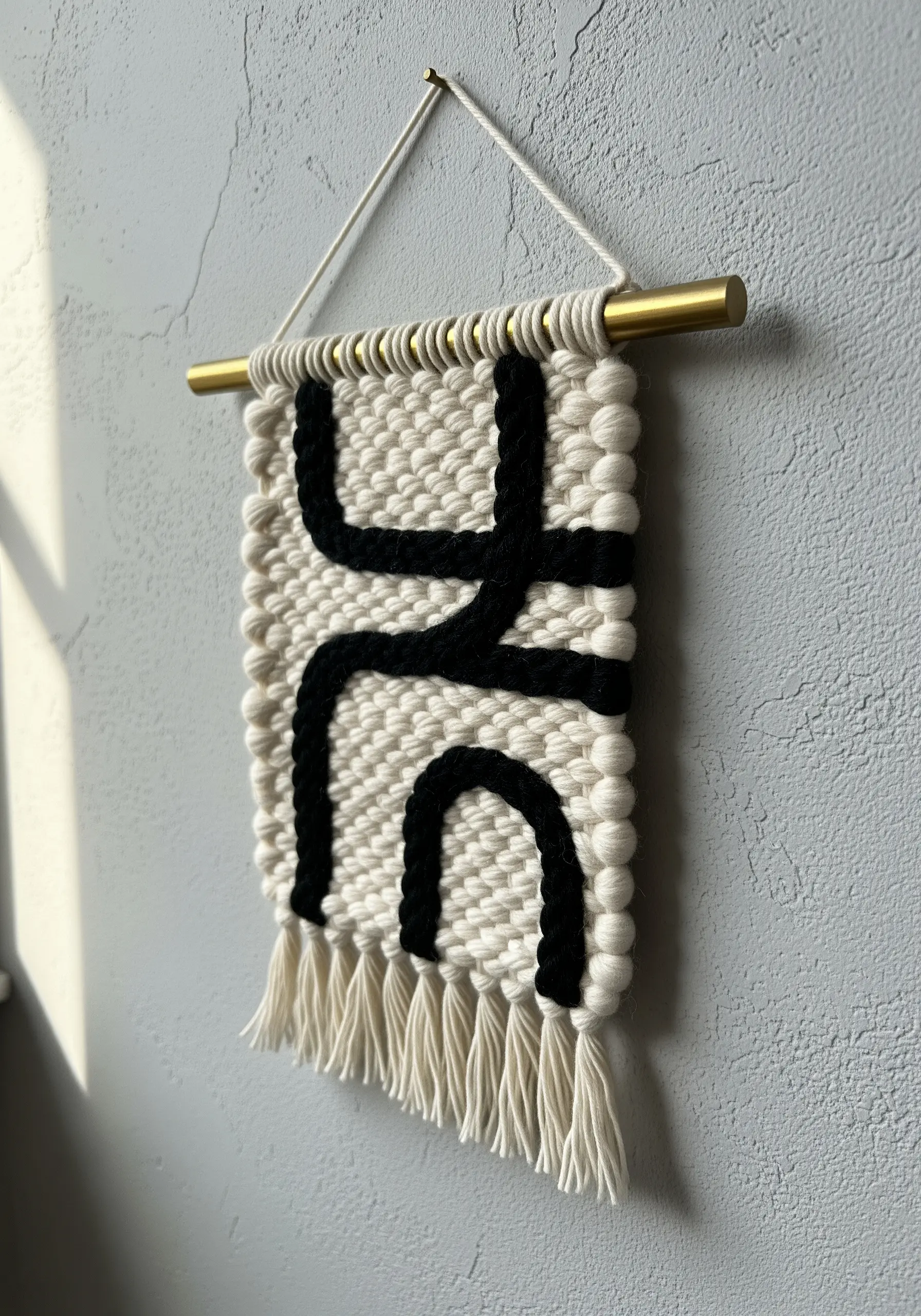 A small woven wall hanging with a black abstract design on a textured cream background.