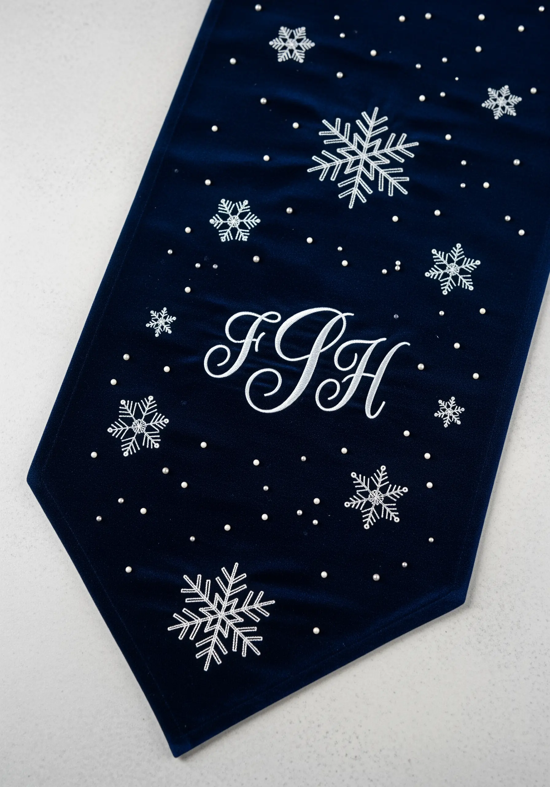 A navy velvet table runner with a white script monogram, snowflakes, and tiny accent beads.