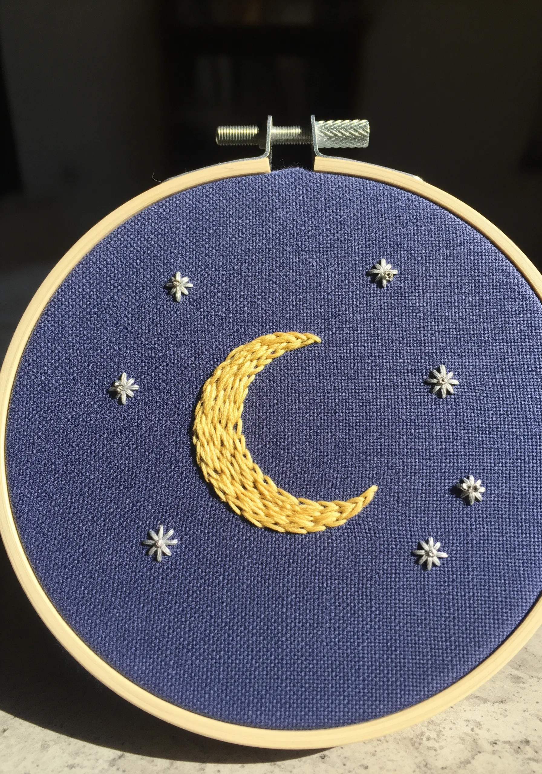 A textured, woven crescent moon in yellow thread surrounded by small white stars on blue fabric.