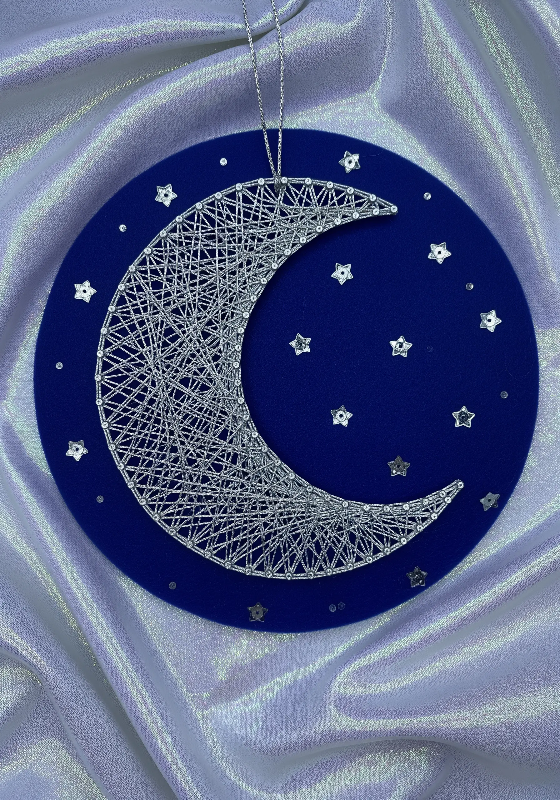 Silver string art crescent moon on a blue background with star sequins.