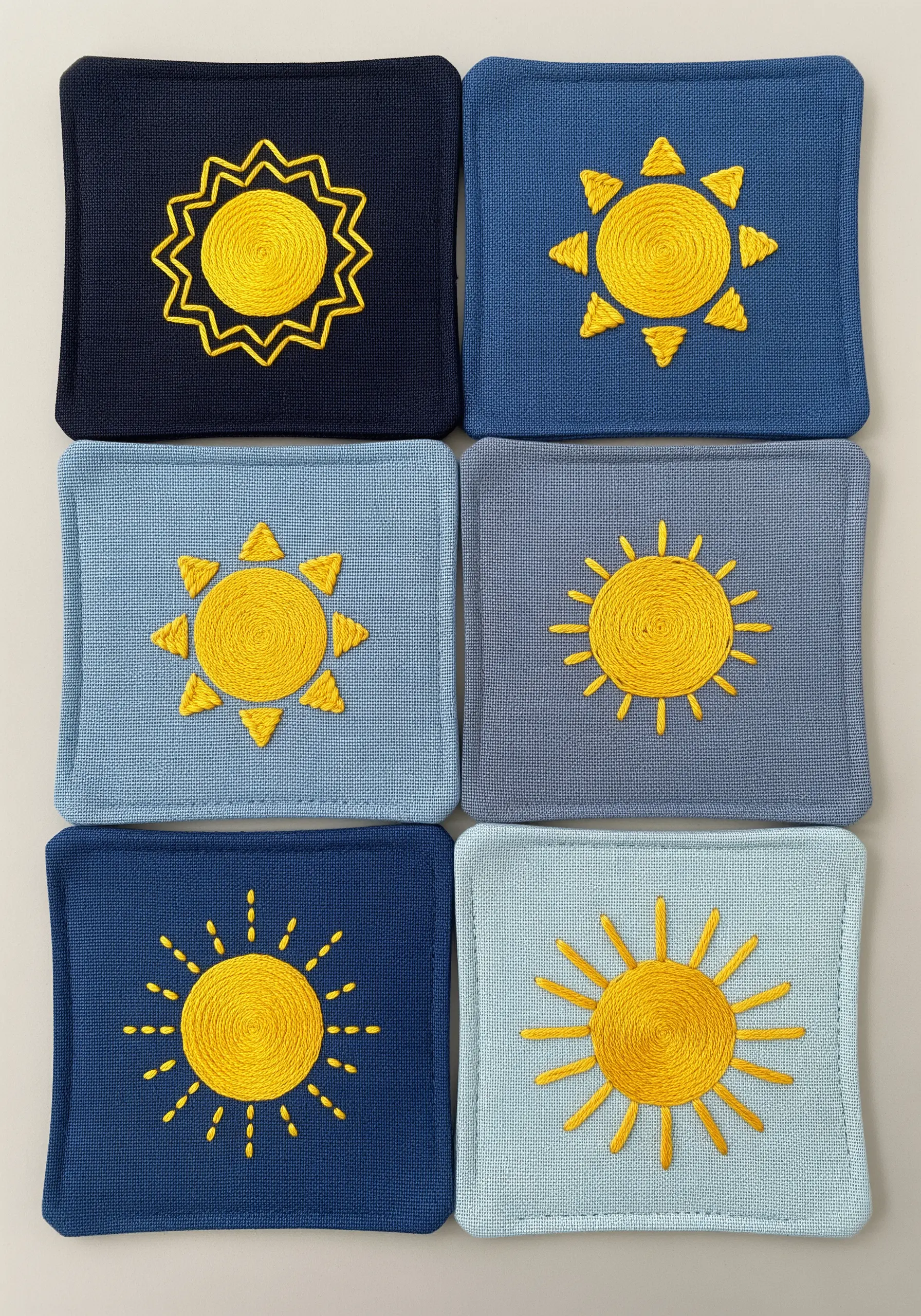 Six yellow embroidered sun motifs on six different blue fabric coasters.