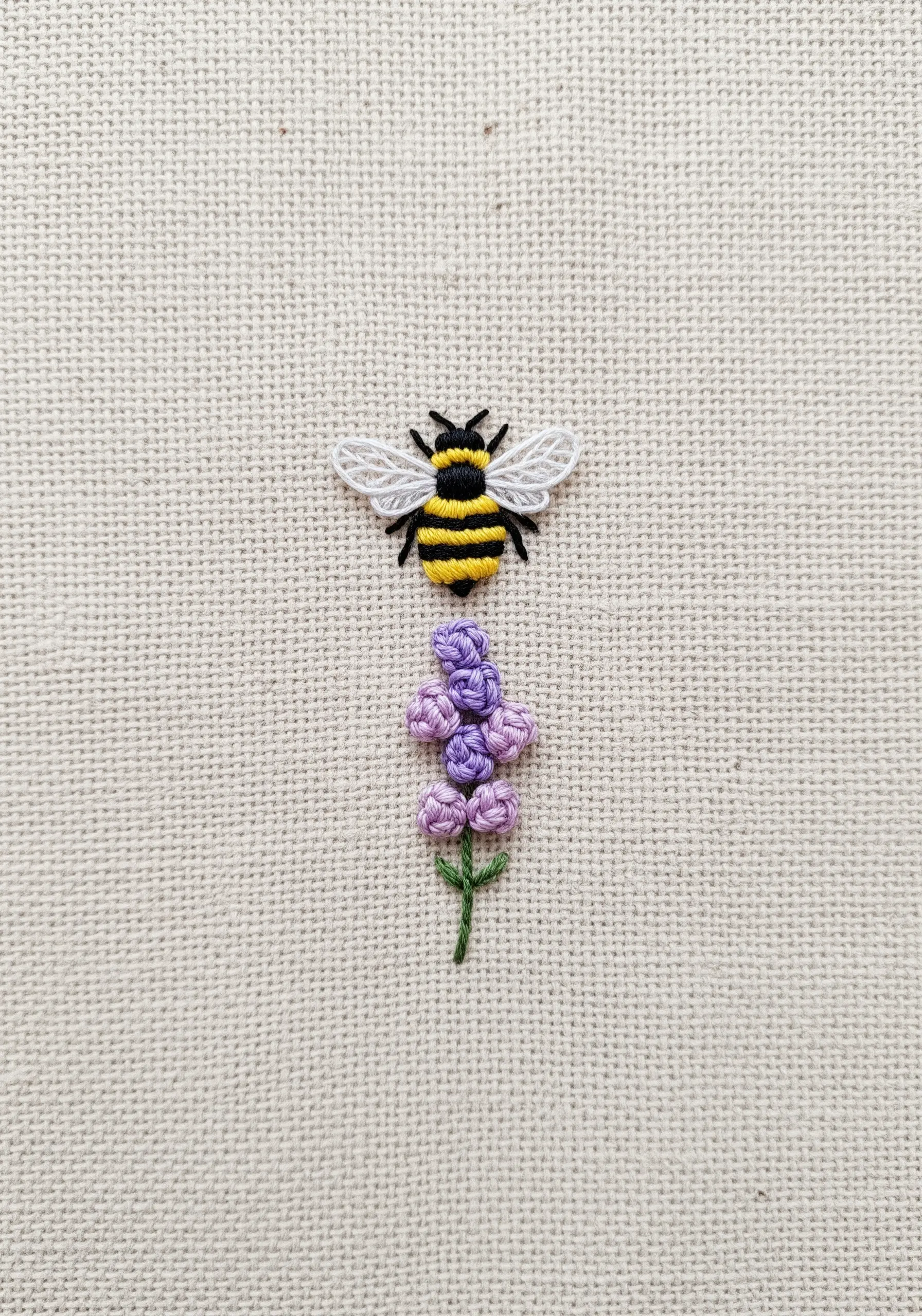A tiny embroidered bee with raised wings flying above a small sprig of lavender.