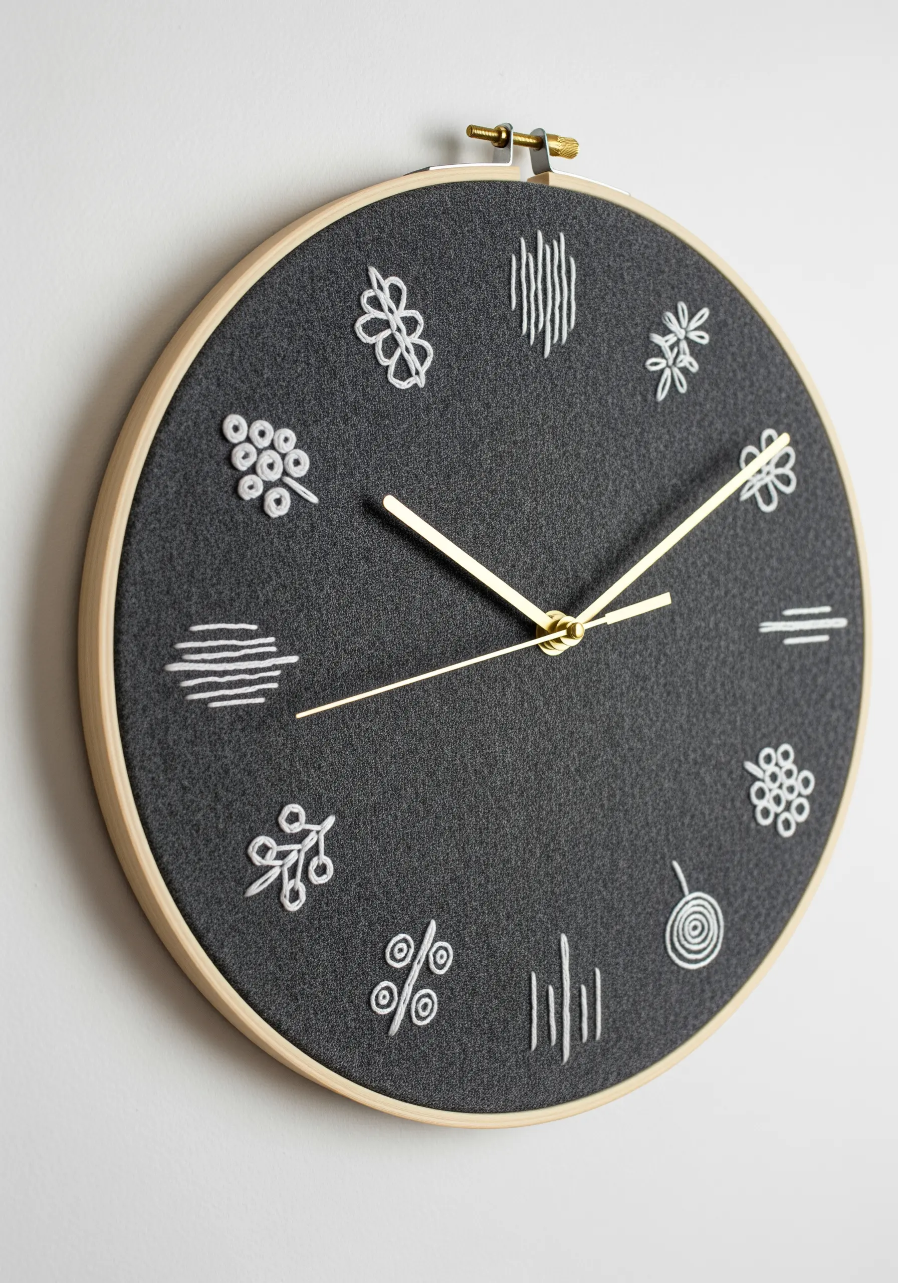 An embroidery hoop clock with a dark grey face and simple white embroidered motifs as hour markers.