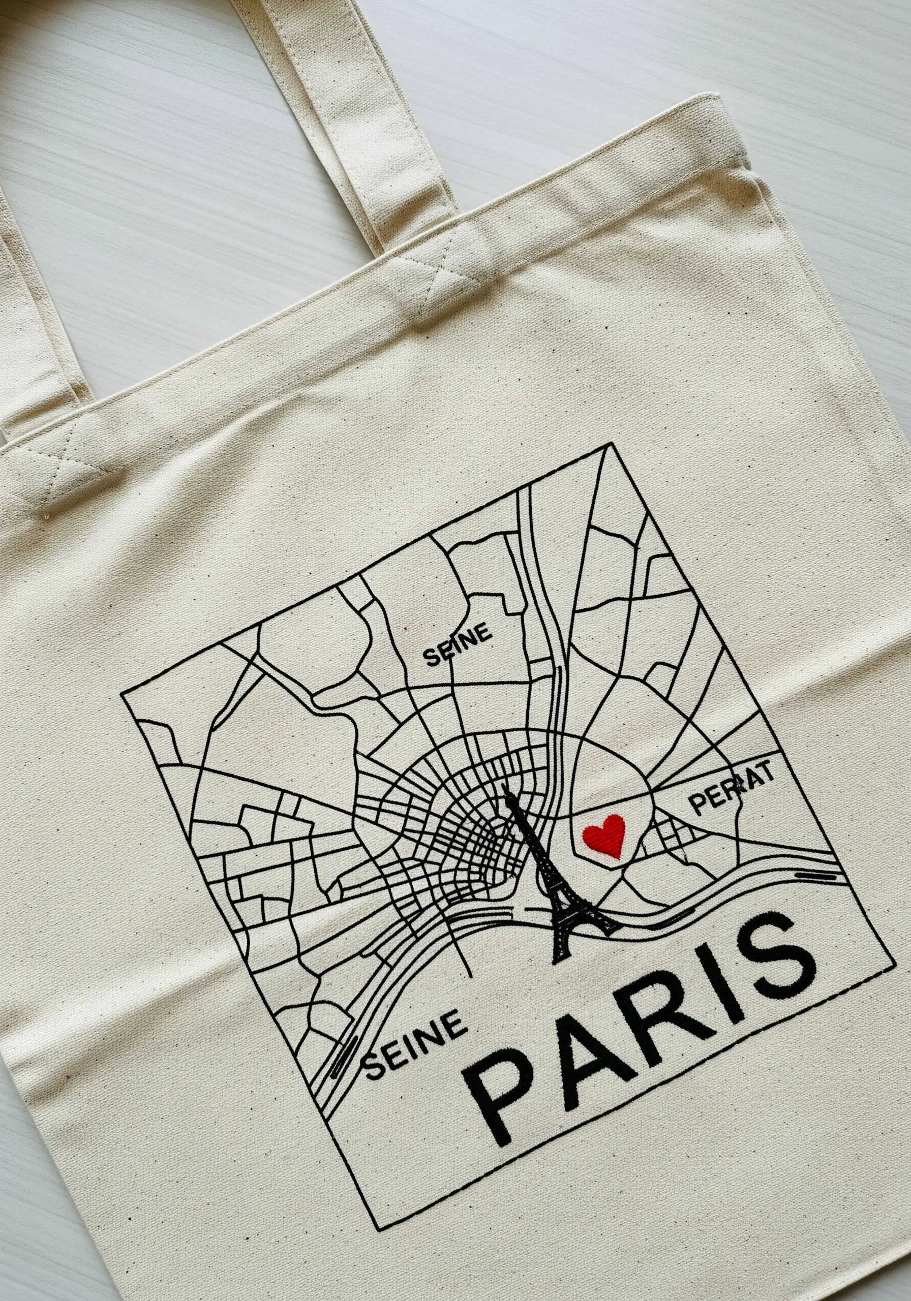 A black line-art map of Paris embroidered on a canvas tote, with a small red heart near the Eiffel Tower.