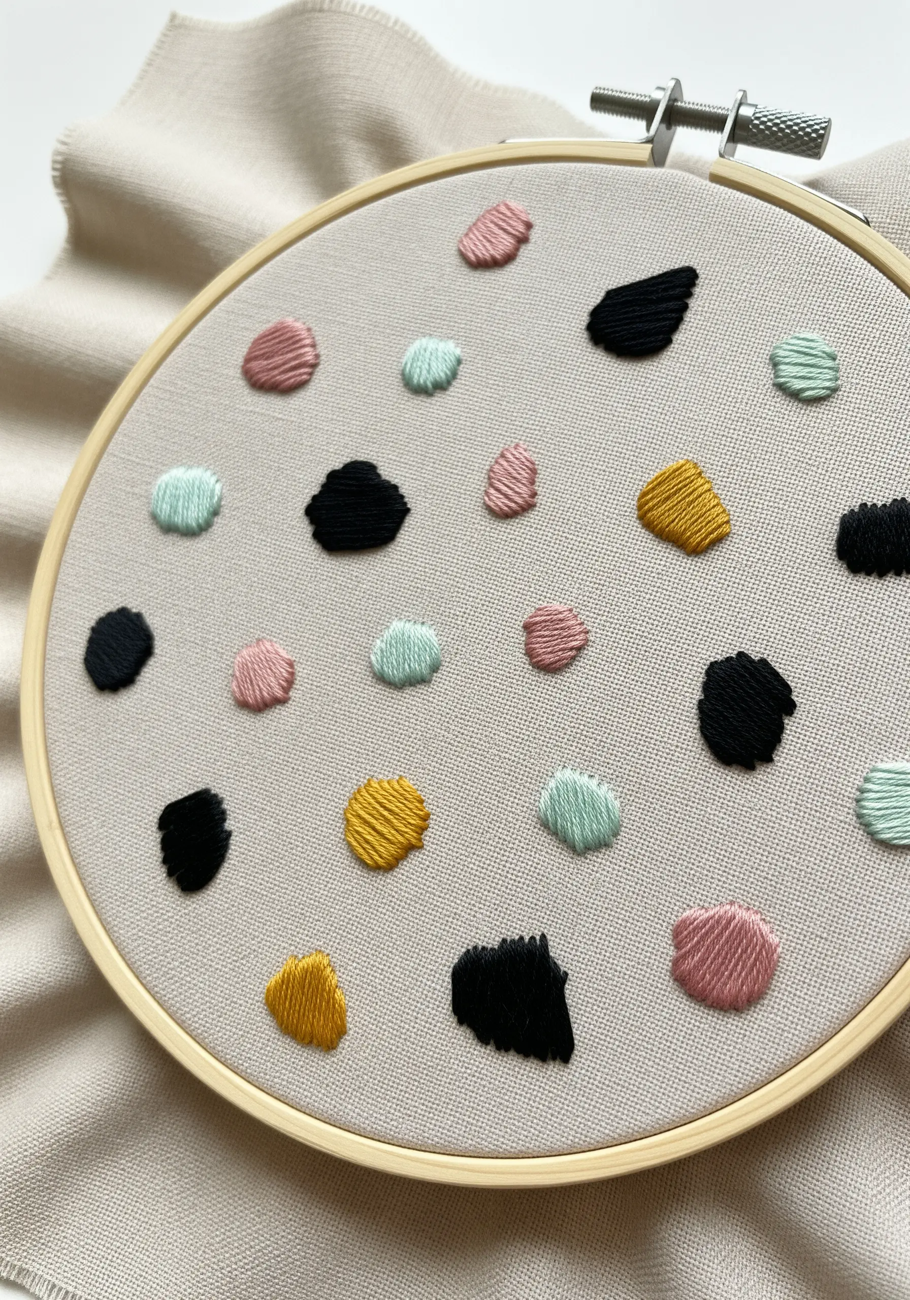 Small, irregular satin stitch shapes in muted colors, scattered like terrazzo on a neutral fabric.
