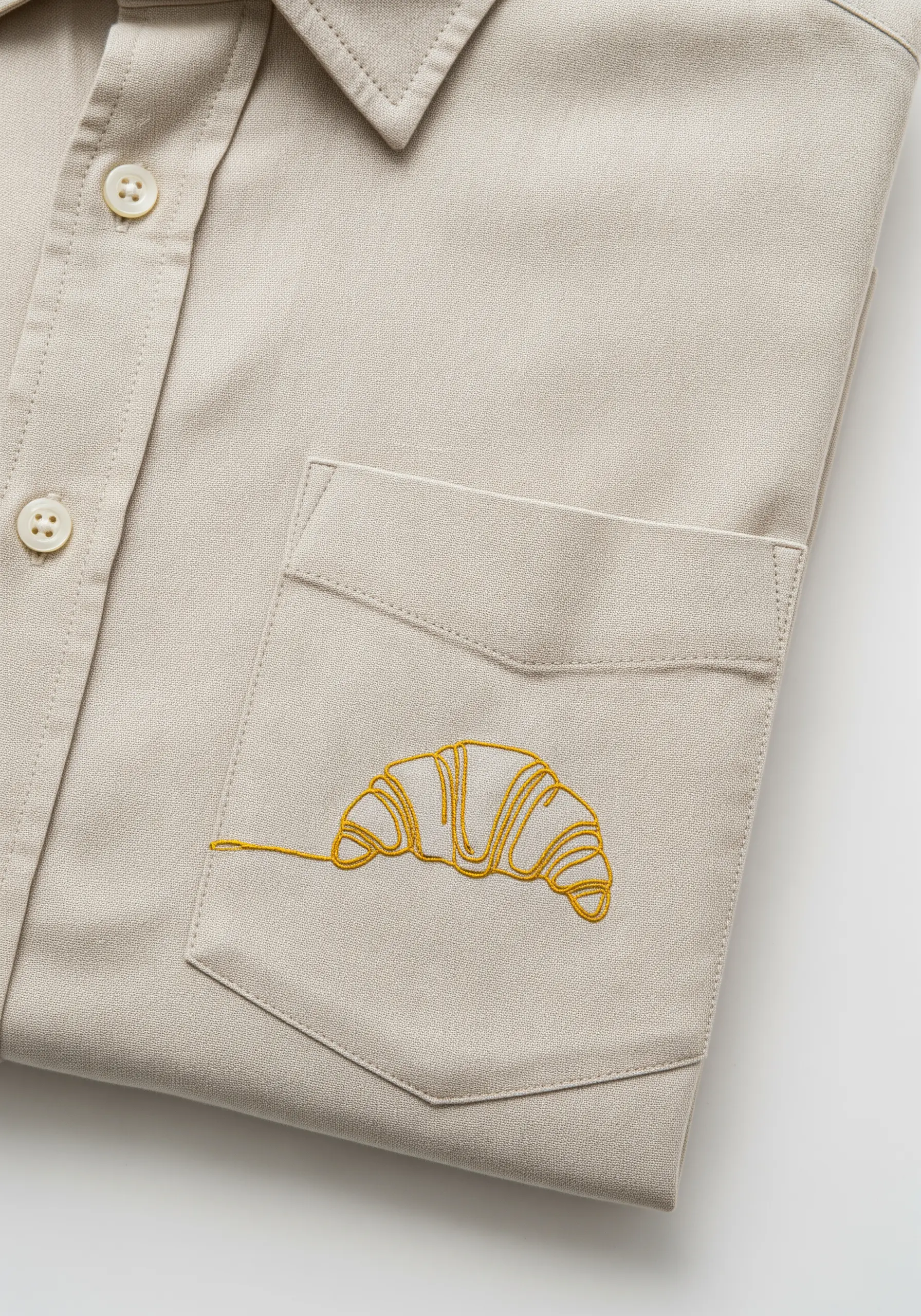 A single-line art croissant embroidered in yellow thread on the pocket of a beige shirt.