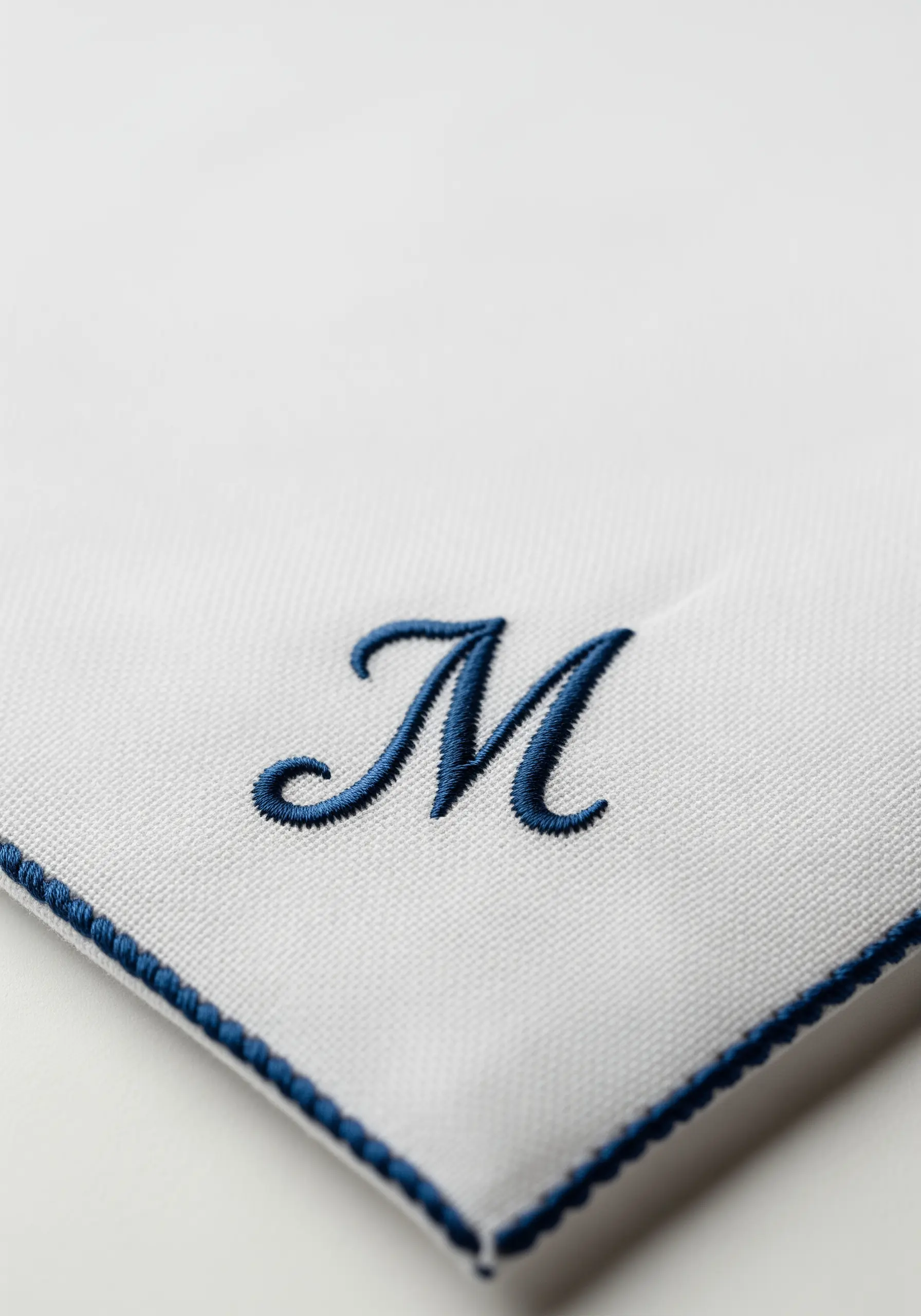 A raised, navy blue embroidered 'M' monogram on the corner of a white handkerchief.