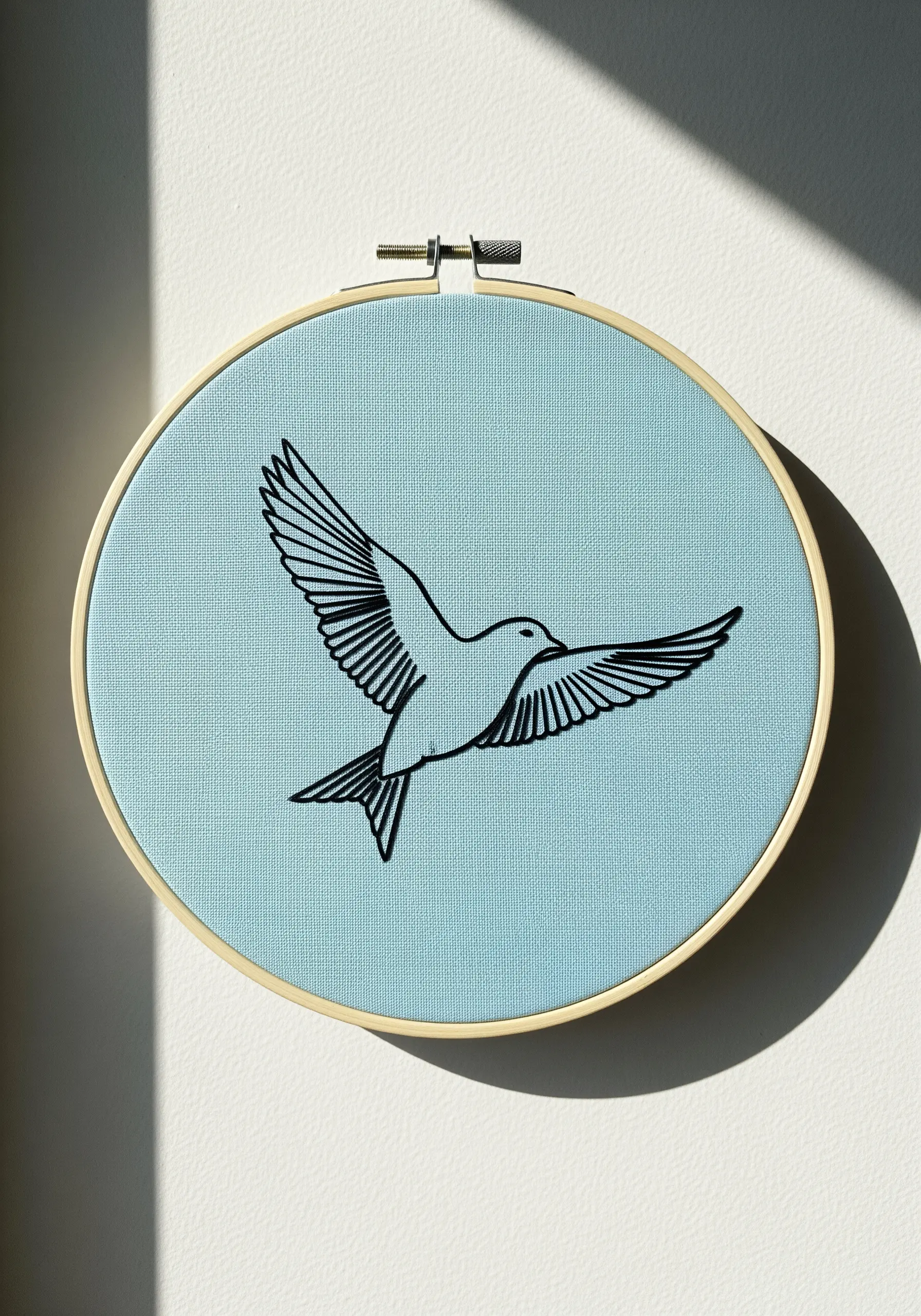 A minimalist black outline of a bird in flight, embroidered on a pale blue fabric hoop.