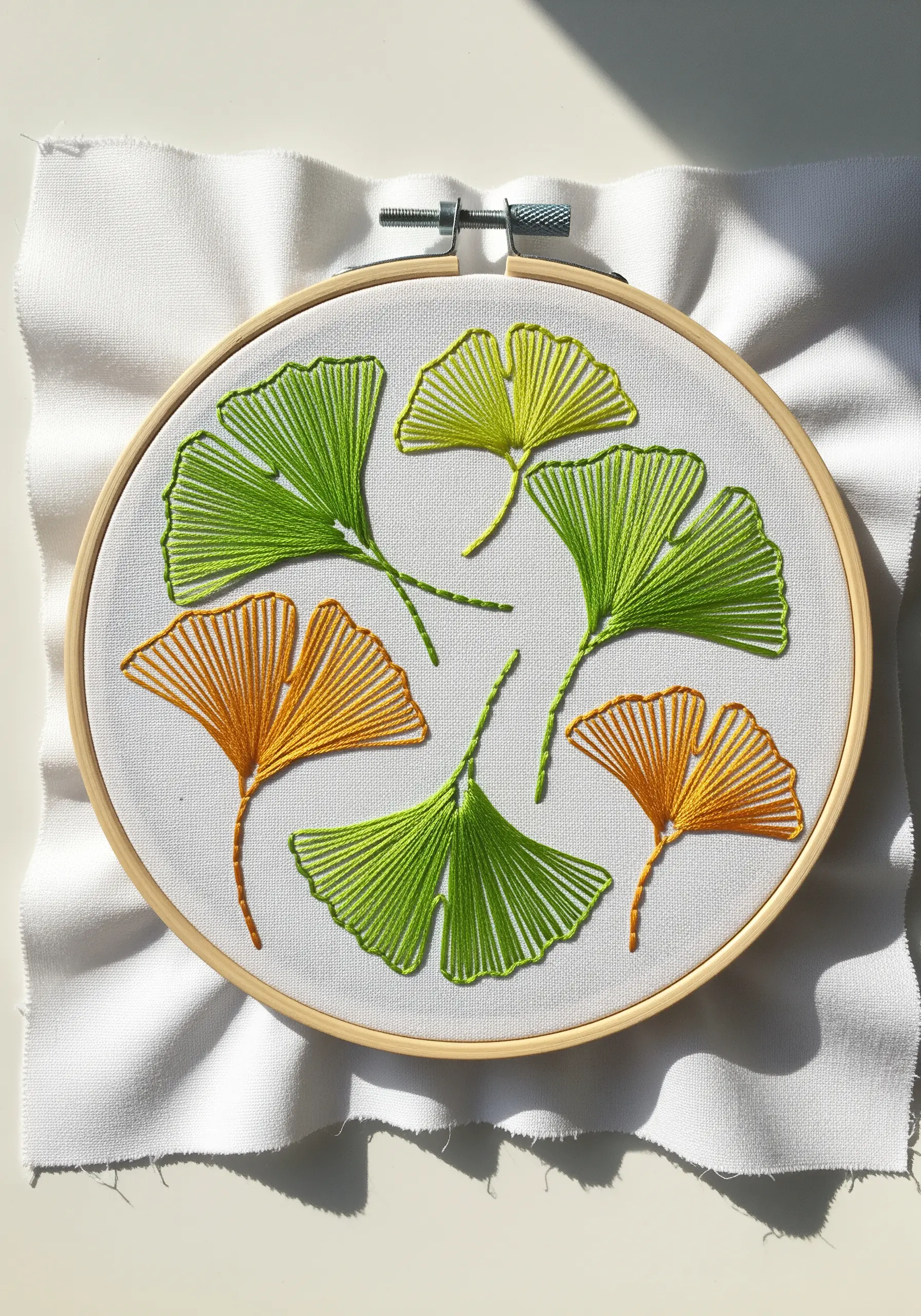Embroidery hoop with minimalist ginkgo leaves in green and yellow satin stitch on white fabric.