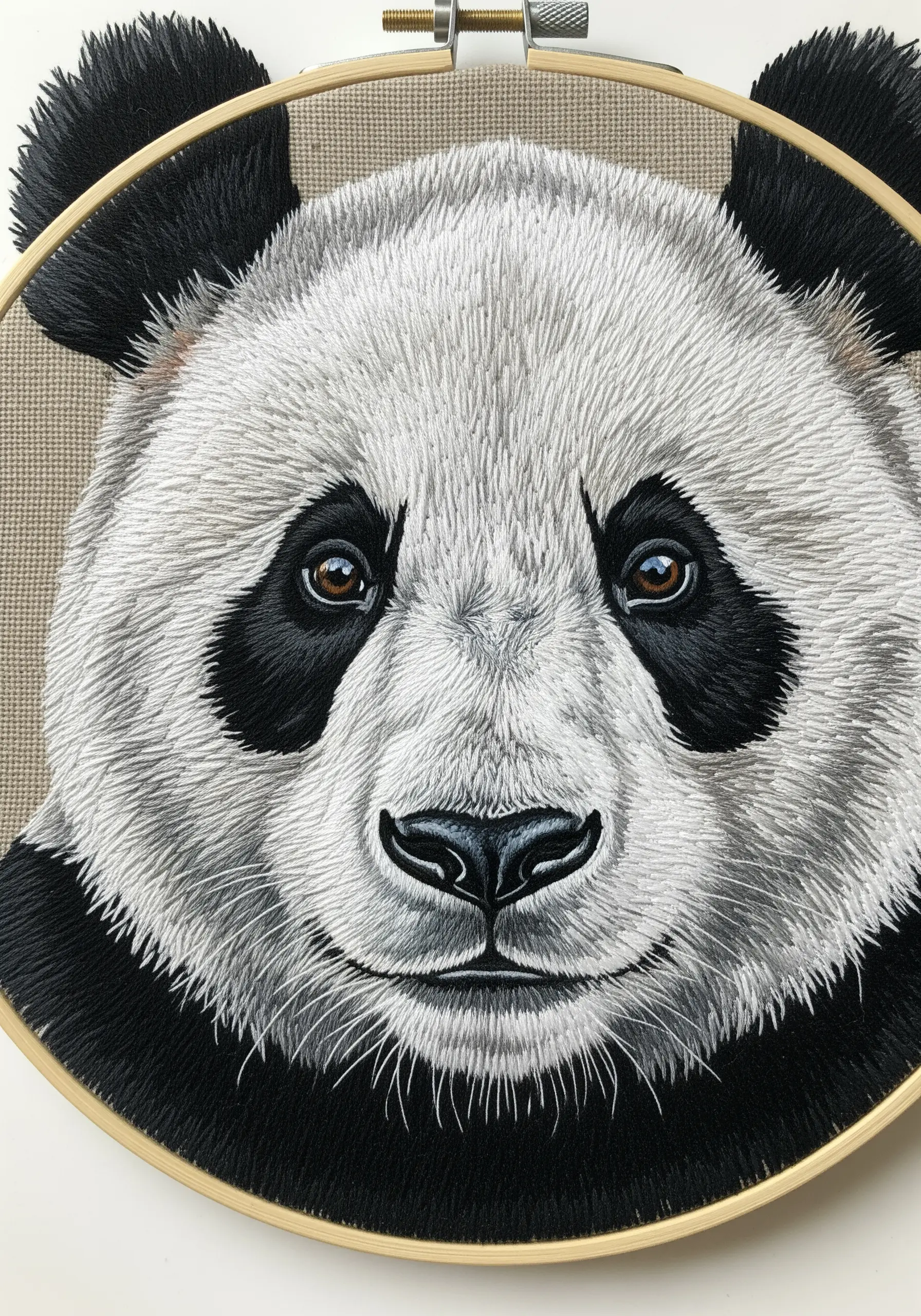 A hyper-realistic, thread-painted portrait of a panda's face in a wooden embroidery hoop.