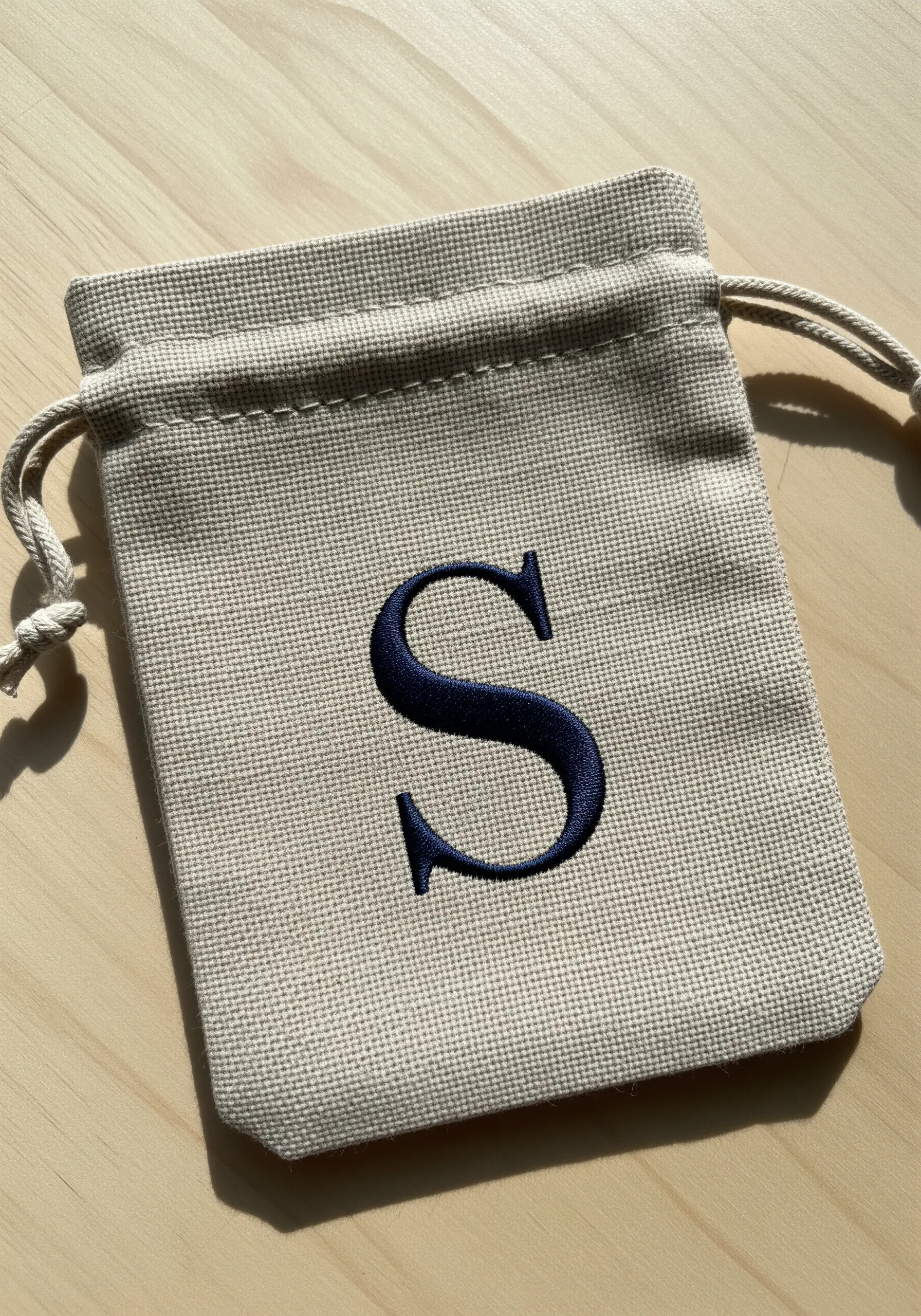 A bold, dark blue embroidered 'S' monogram using a padded satin stitch on a canvas drawstring pouch.