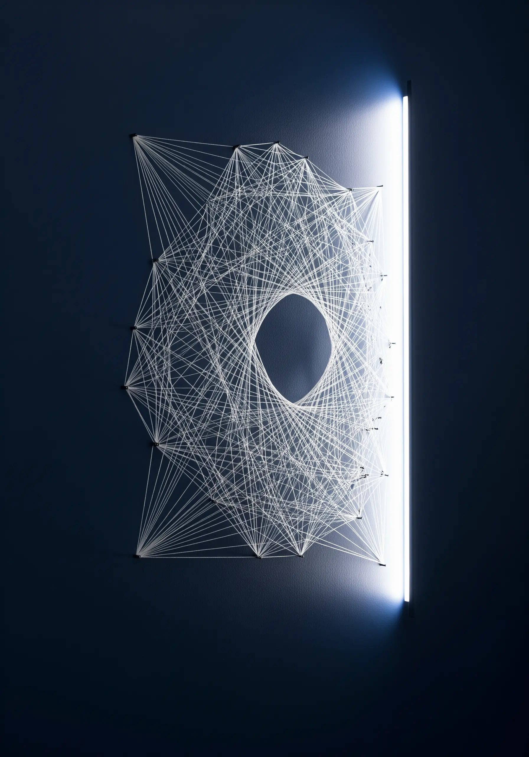 Geometric string art on a dark wall, illuminated from the side by a glowing light bar.
