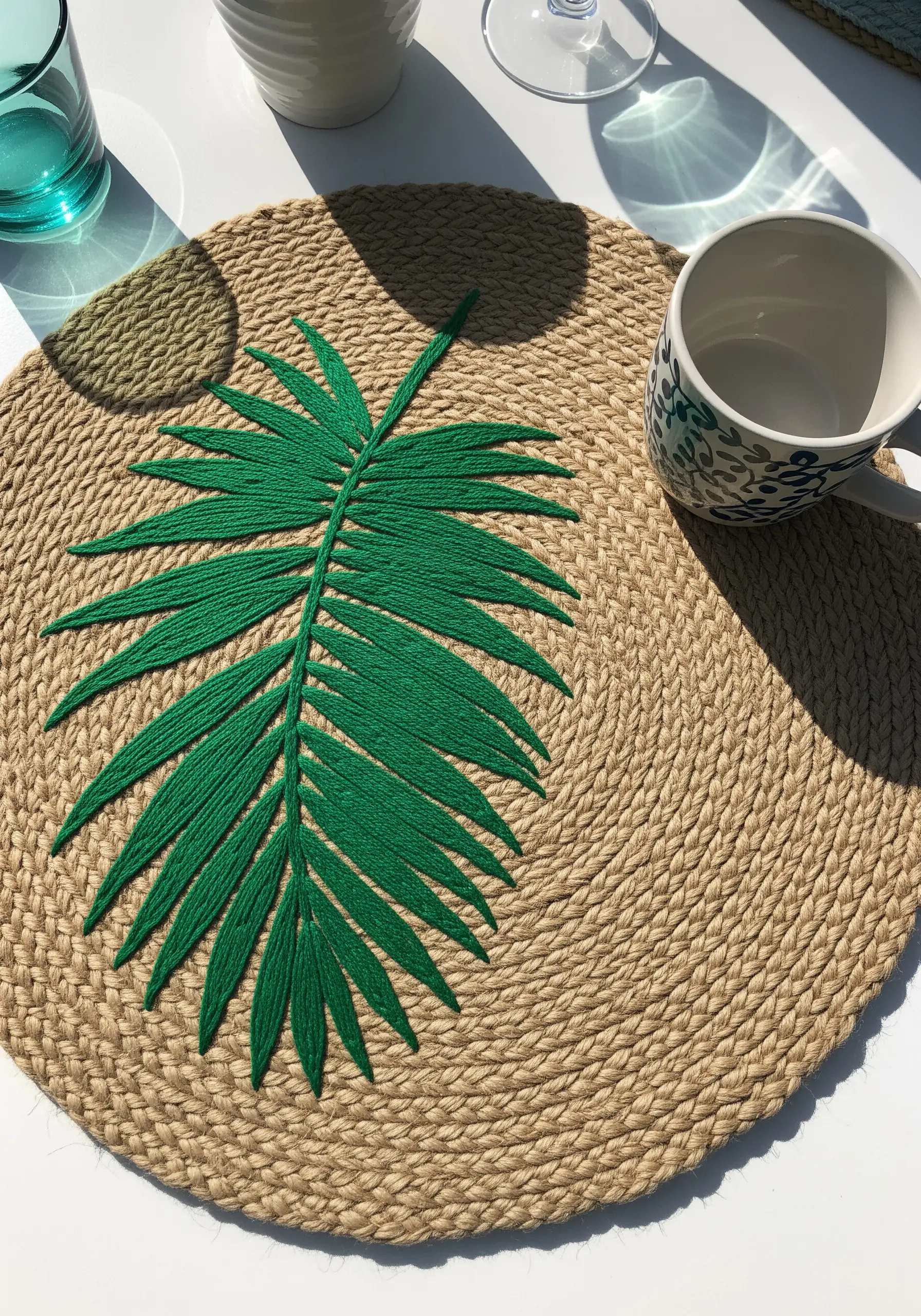 A large palm leaf embroidered with thick green yarn onto a round, woven jute placemat.