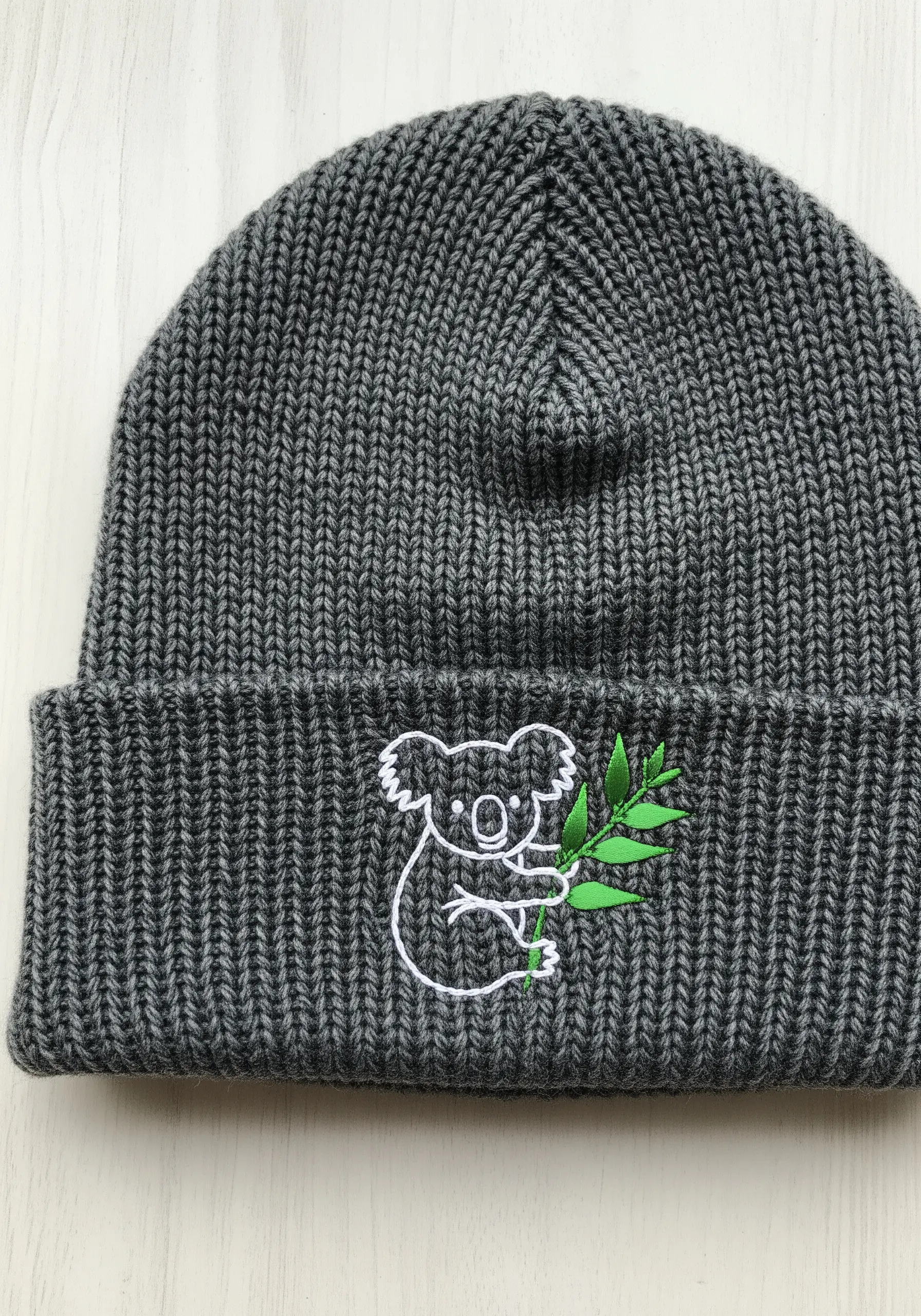 A simple white line-art koala with green satin-stitch leaves on a grey ribbed beanie.