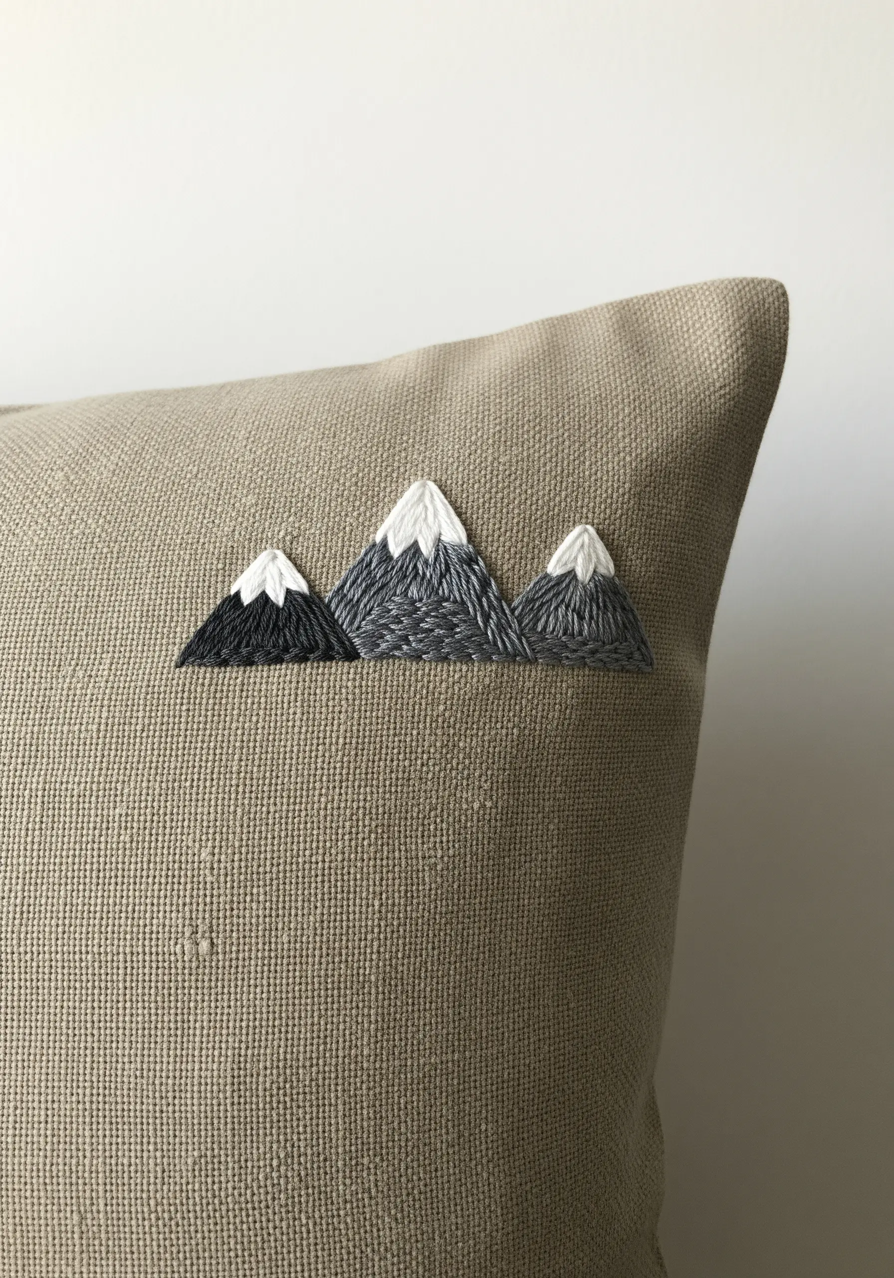Three small embroidered mountains with white peaks on the corner of a textured beige pillowcase.