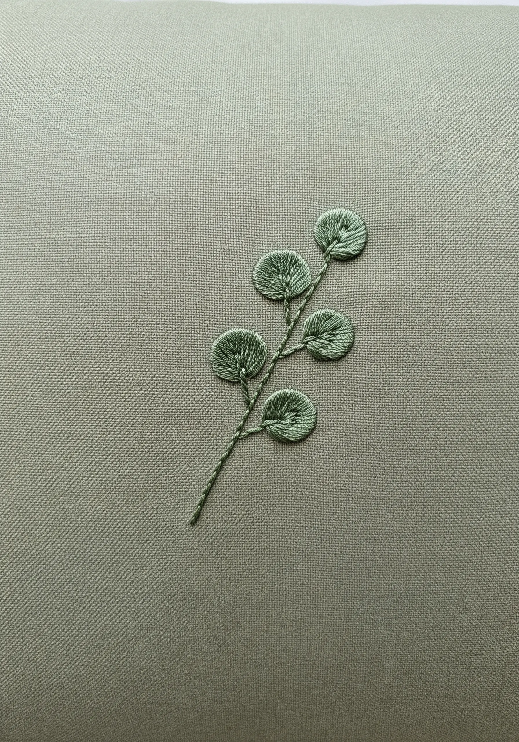 A single sprig of eucalyptus with round leaves embroidered in green thread on a sage green pillowcase.