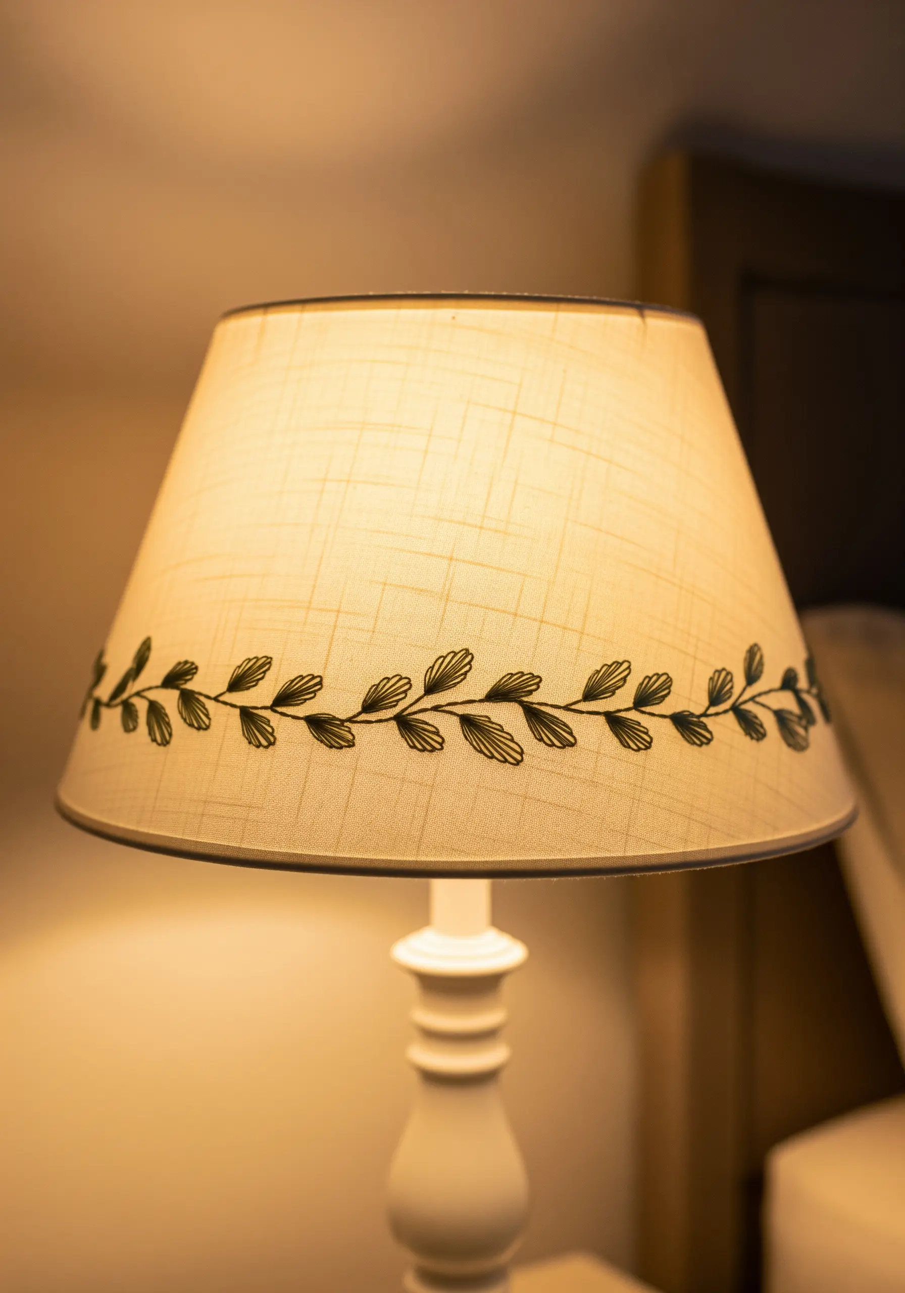A simple black leaf vine embroidered around the middle of a lit fabric lampshade.