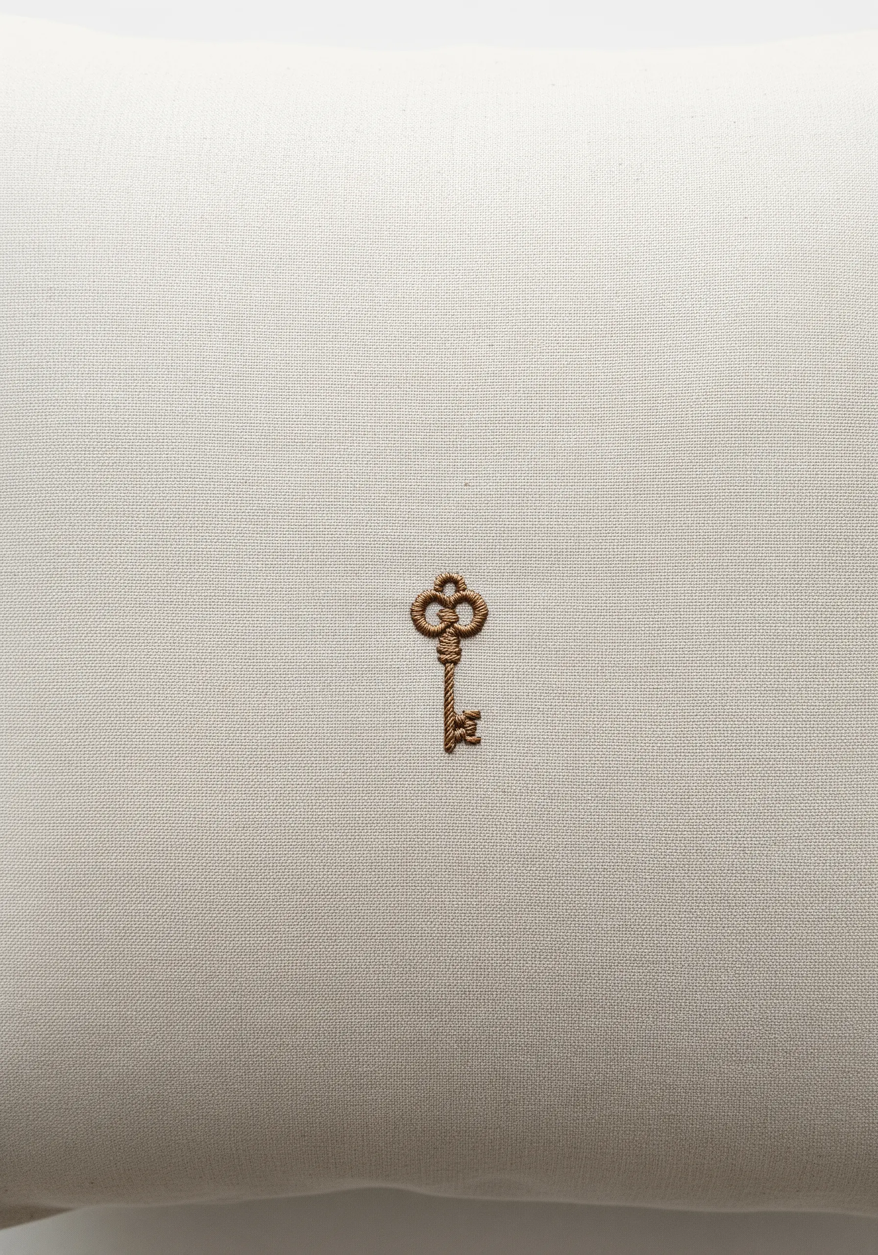 A small, ornate antique-style key embroidered in brown thread on a light beige pillowcase.