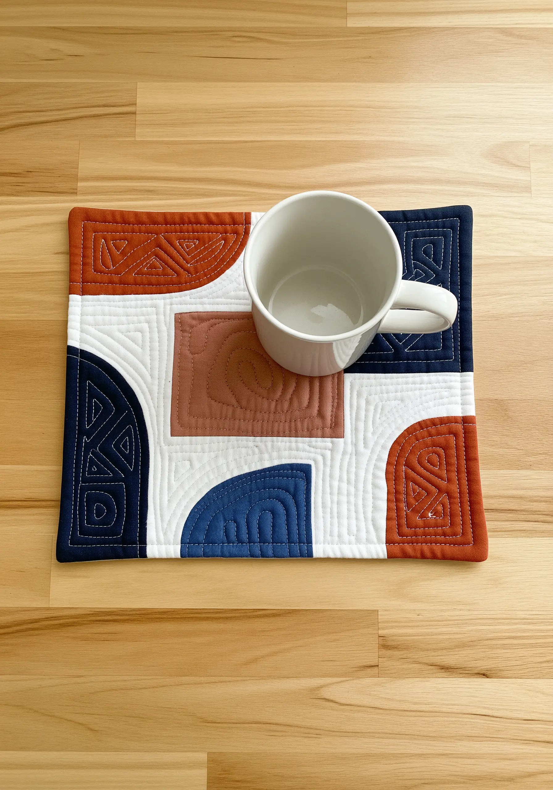 A modern quilted mug rug with abstract shapes in rust, blue, and white, accented with hand stitching.