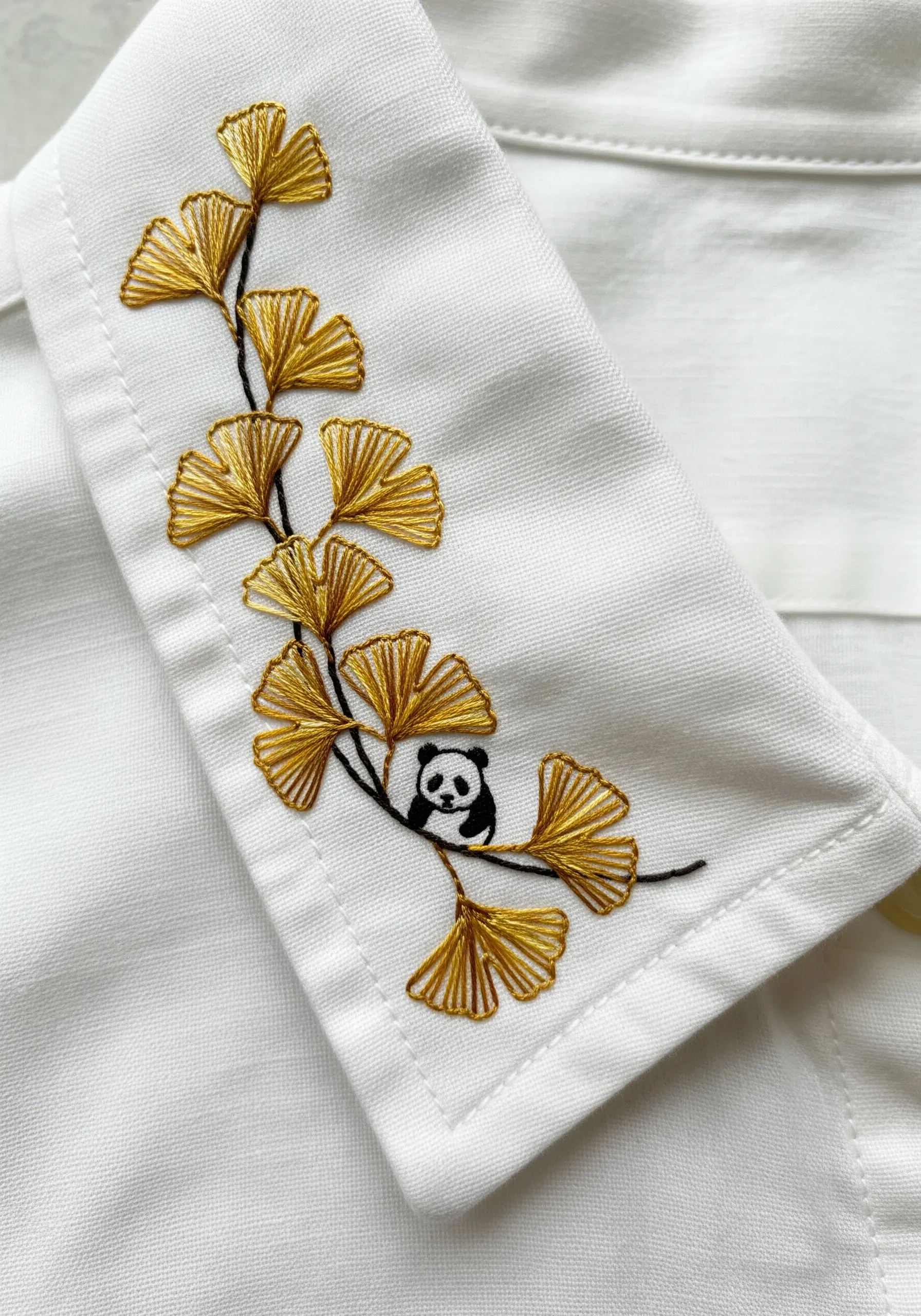 A tiny panda climbing a branch of golden ginkgo leaves embroidered on a white shirt collar.