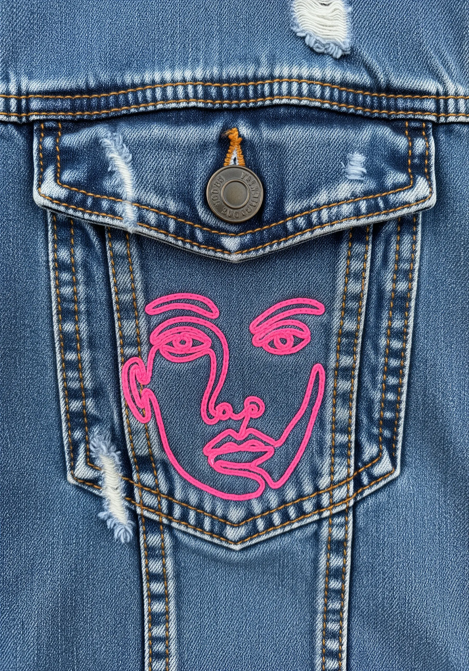 Bright neon pink line-art face embroidered on the pocket of a distressed denim jacket.