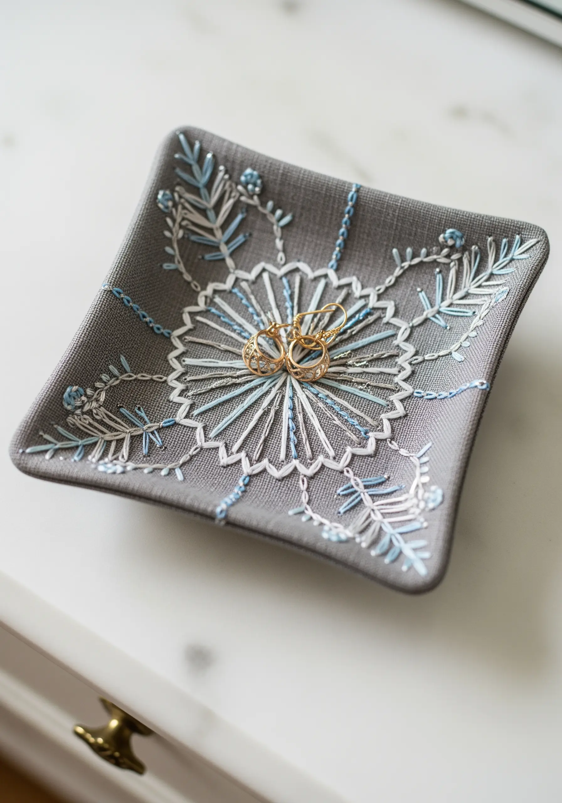 A square, grey linen dish with a white and blue embroidered mandala, holding a pair of rings.