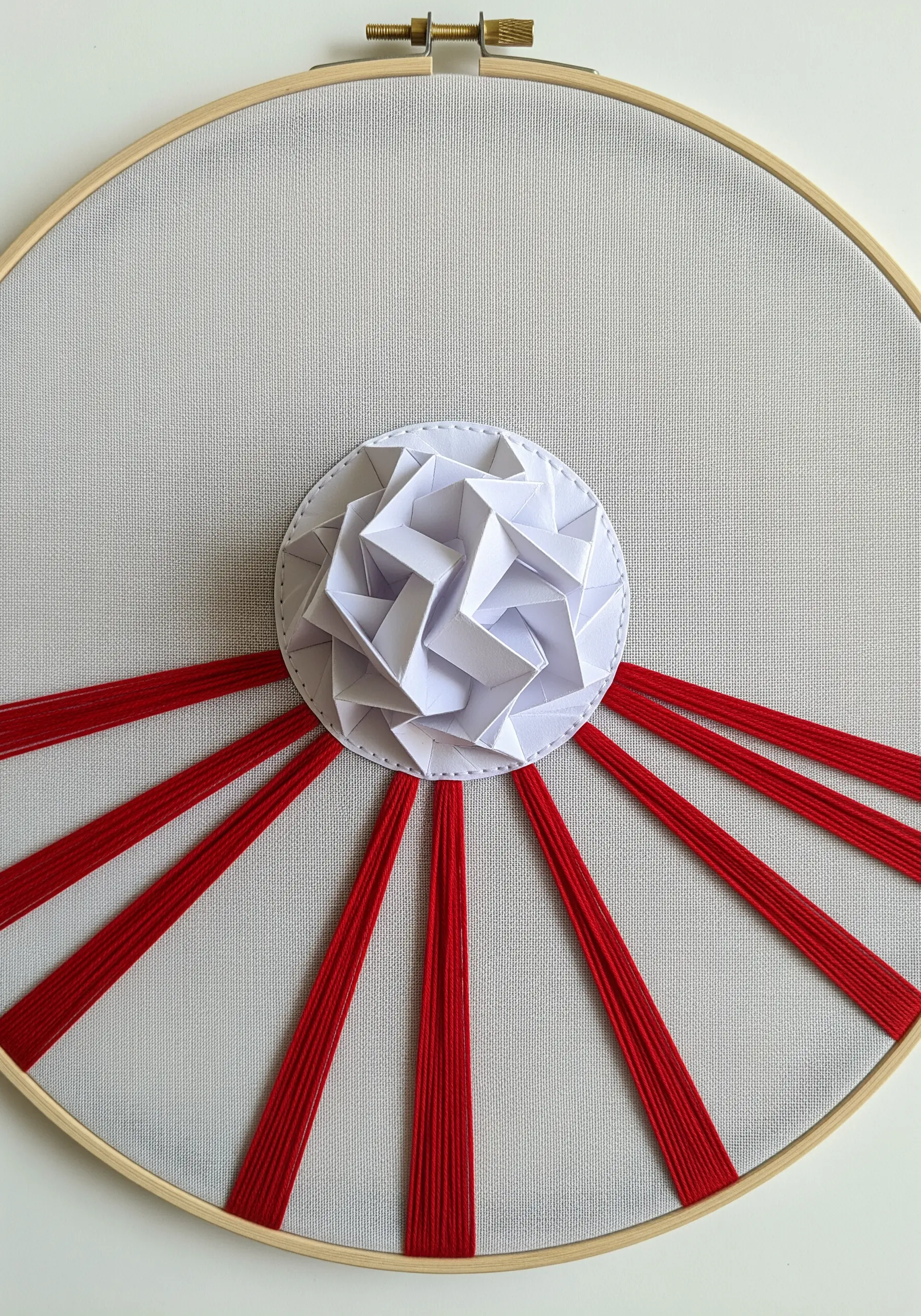 A white geometric origami circle at the center of a hoop, with bold red threads radiating outwards.