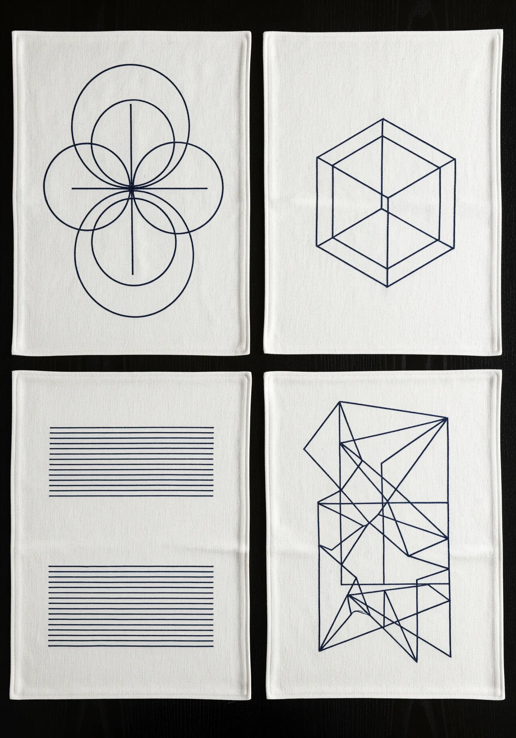 Four white napkins with different modern geometric designs embroidered in dark blue thread.