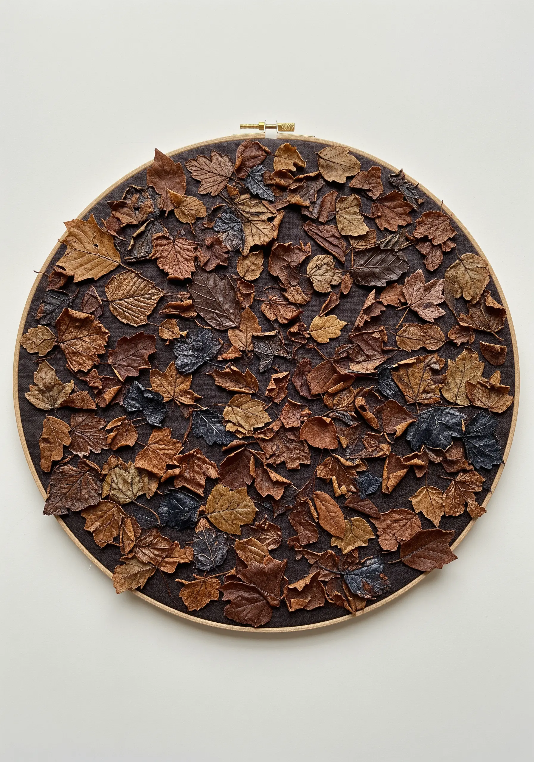 An embroidery hoop filled with a dense collage of real, dried autumn leaves on a dark brown fabric.