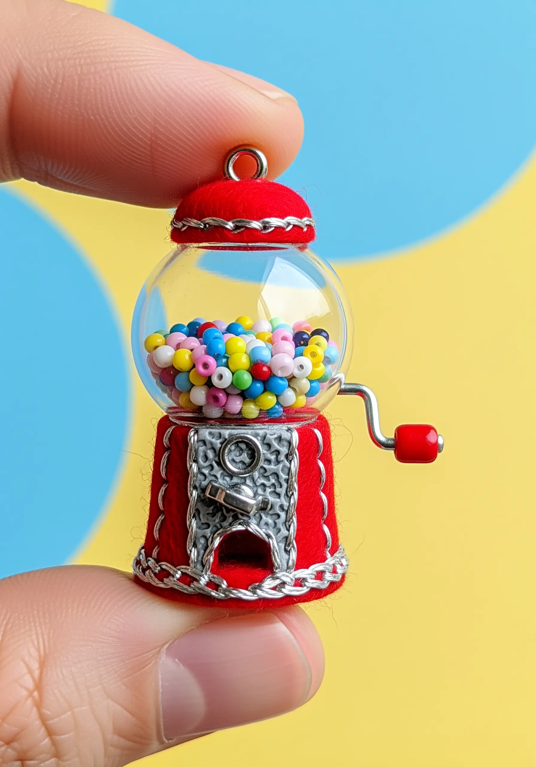 A miniature gumball machine charm made of red felt, silver thread, and a glass orb full of beads.