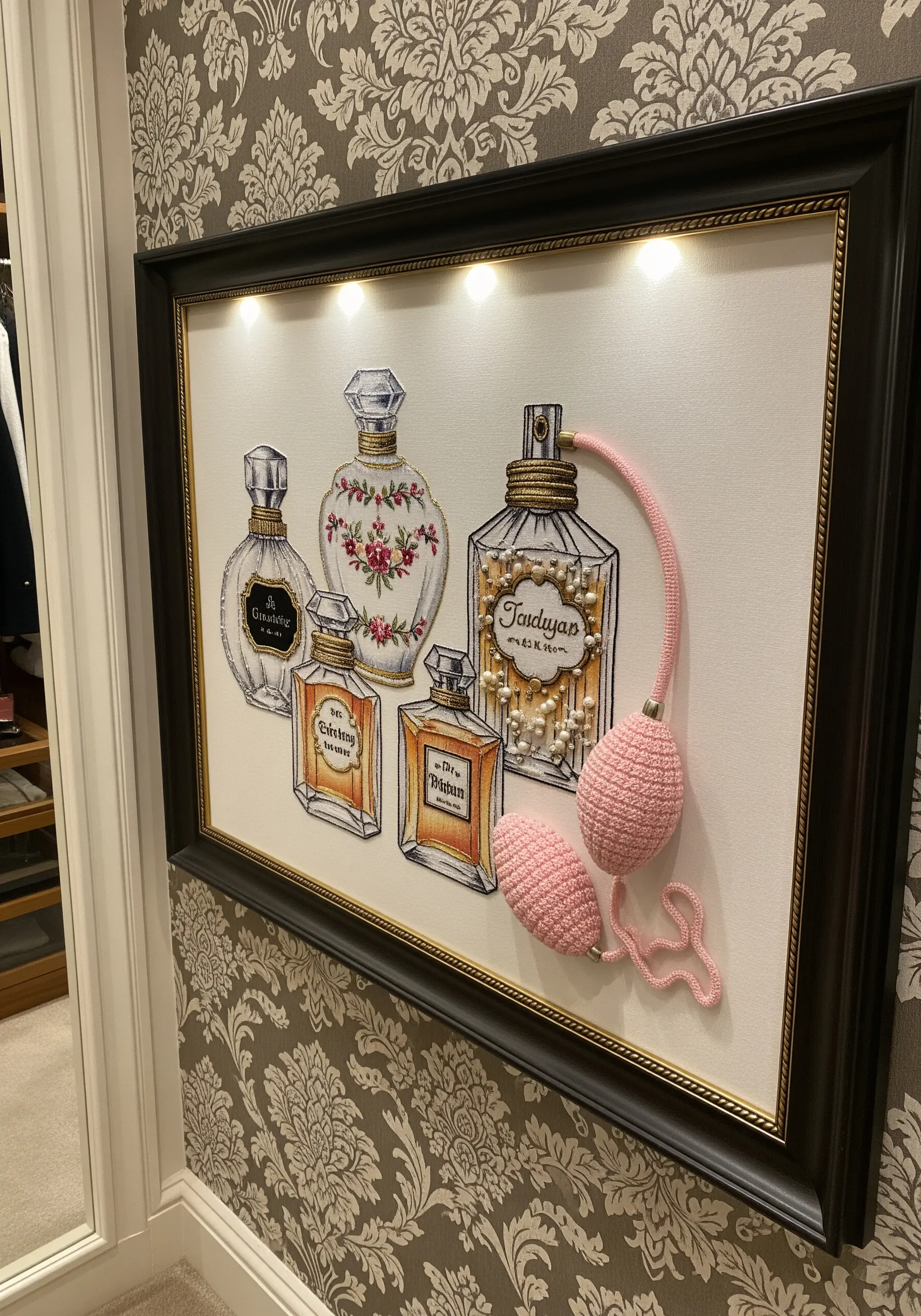 A framed embroidery of vintage perfume bottles with a 3D crocheted pink atomizer attached