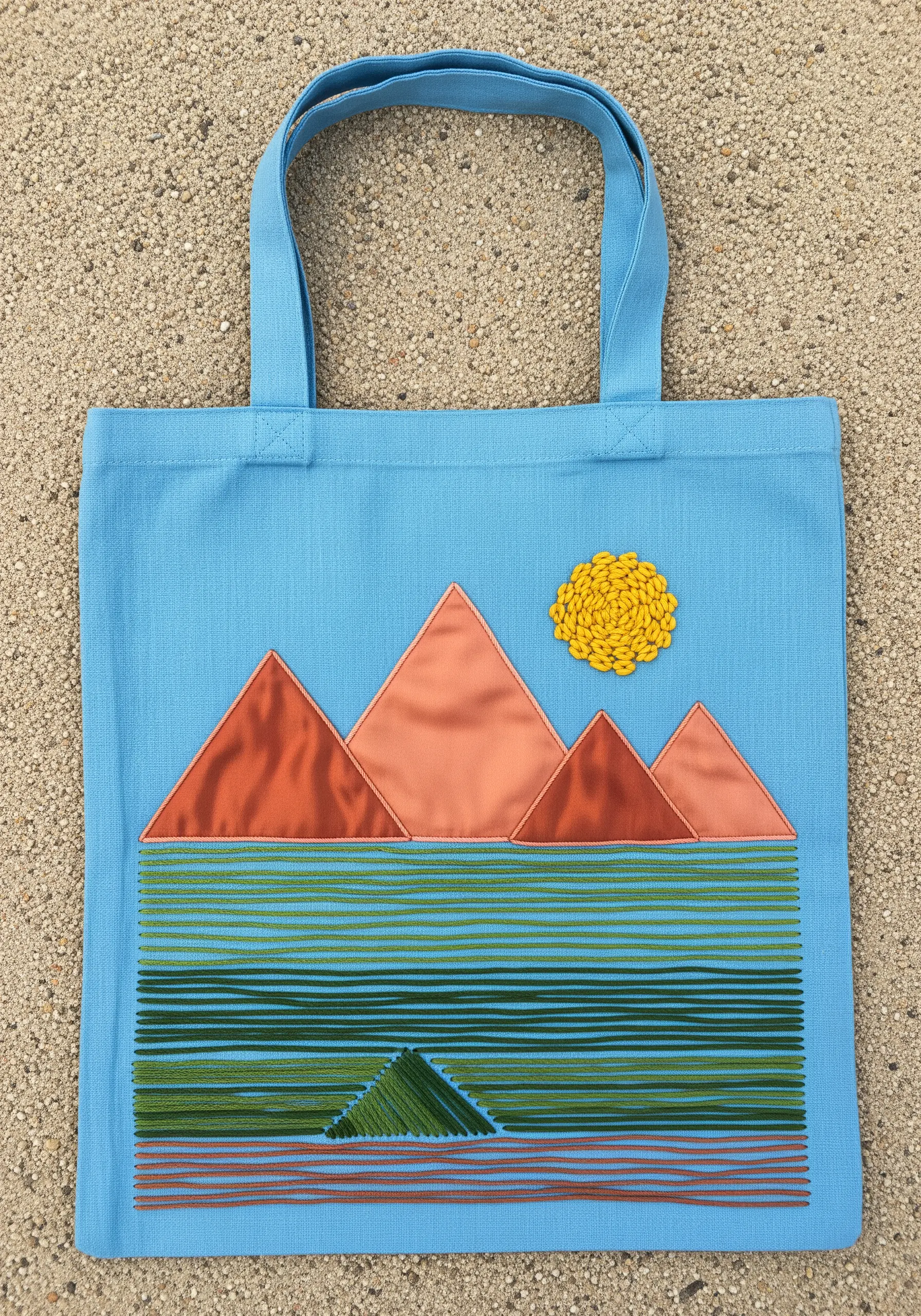 Geometric landscape on a blue tote with appliqué mountains and stitched water and sun.