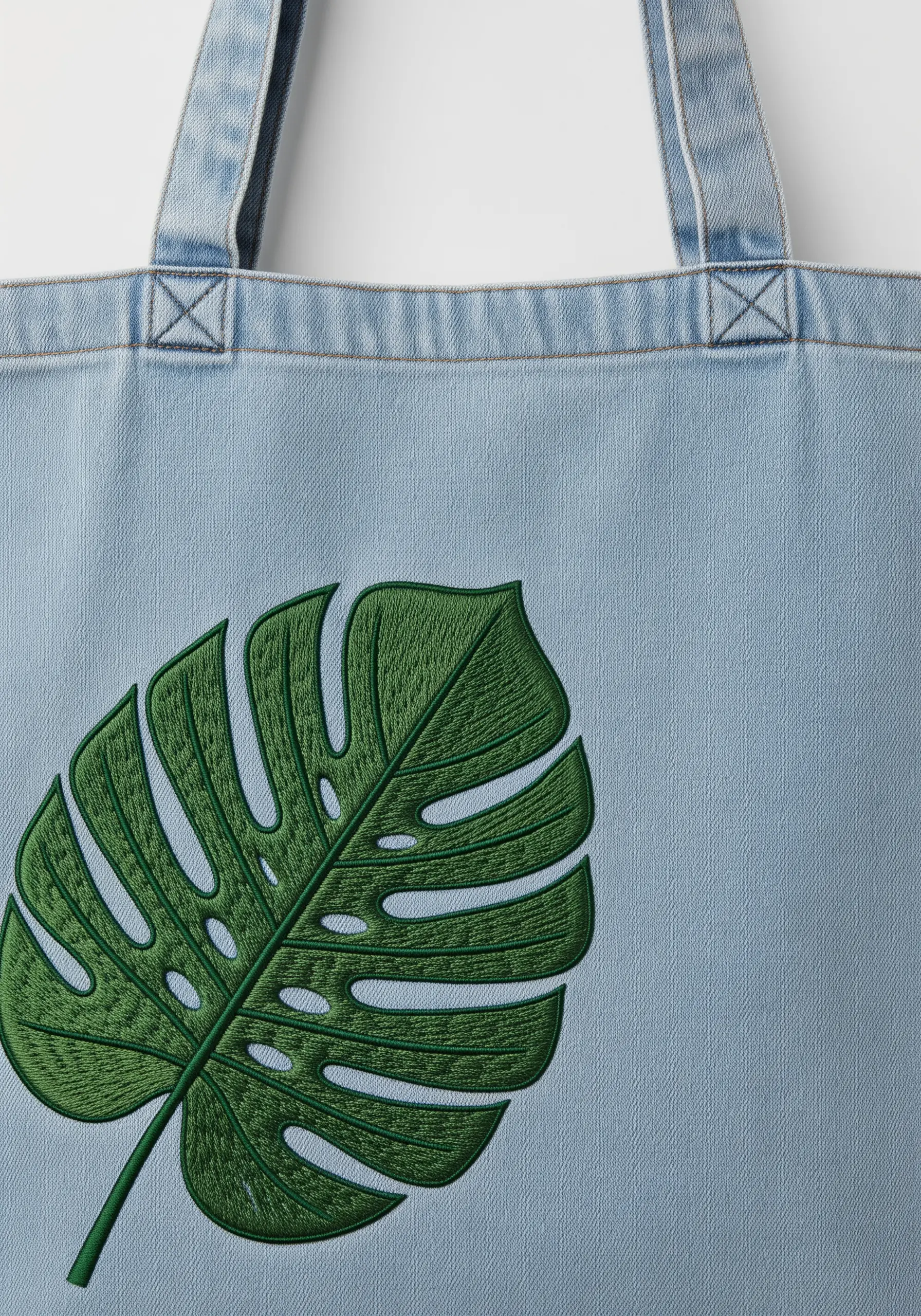 A large, vibrant green Monstera leaf embroidered on a light-wash denim tote.