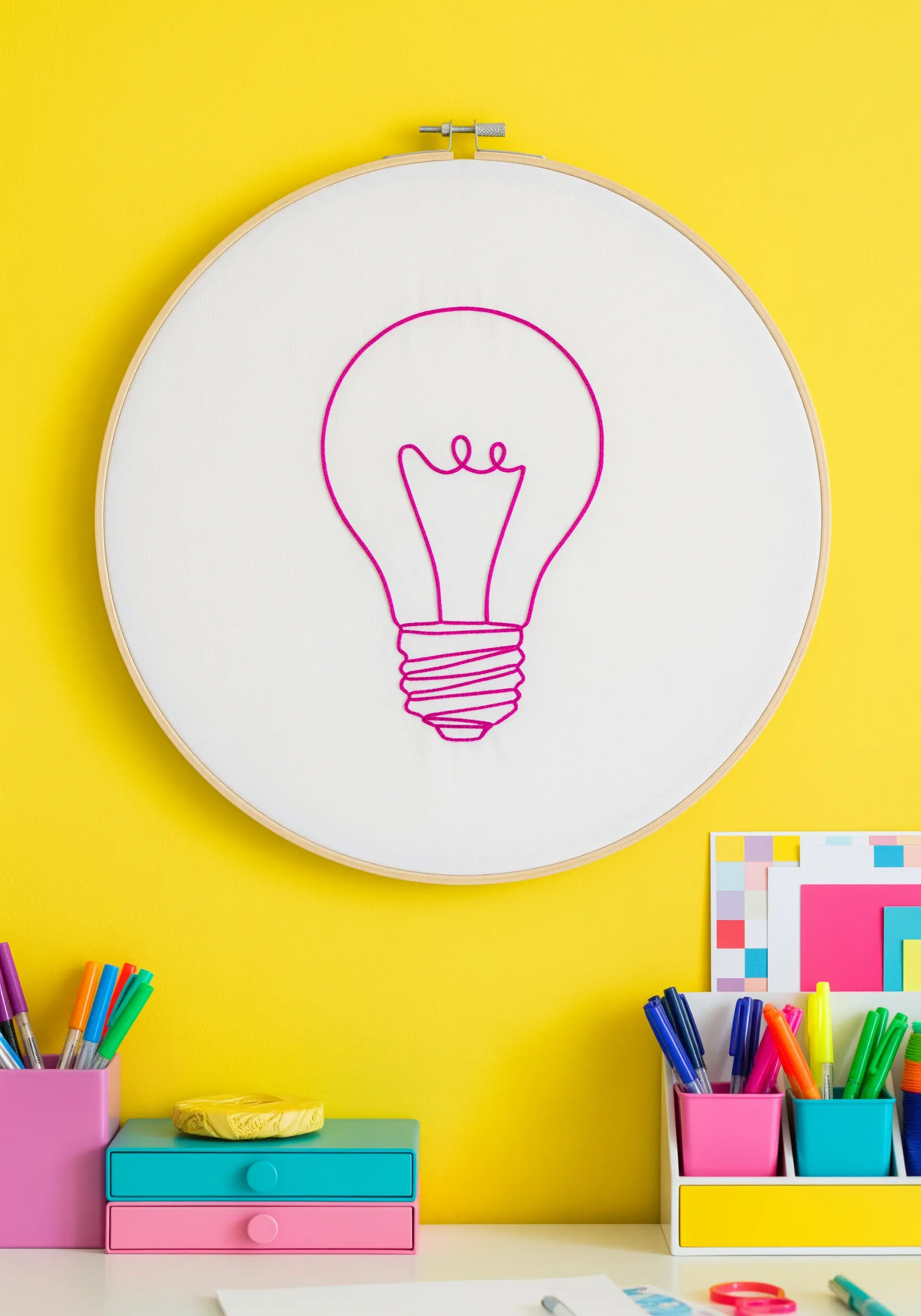 A bright pink chain stitch outline of a lightbulb on white fabric in a large hoop.